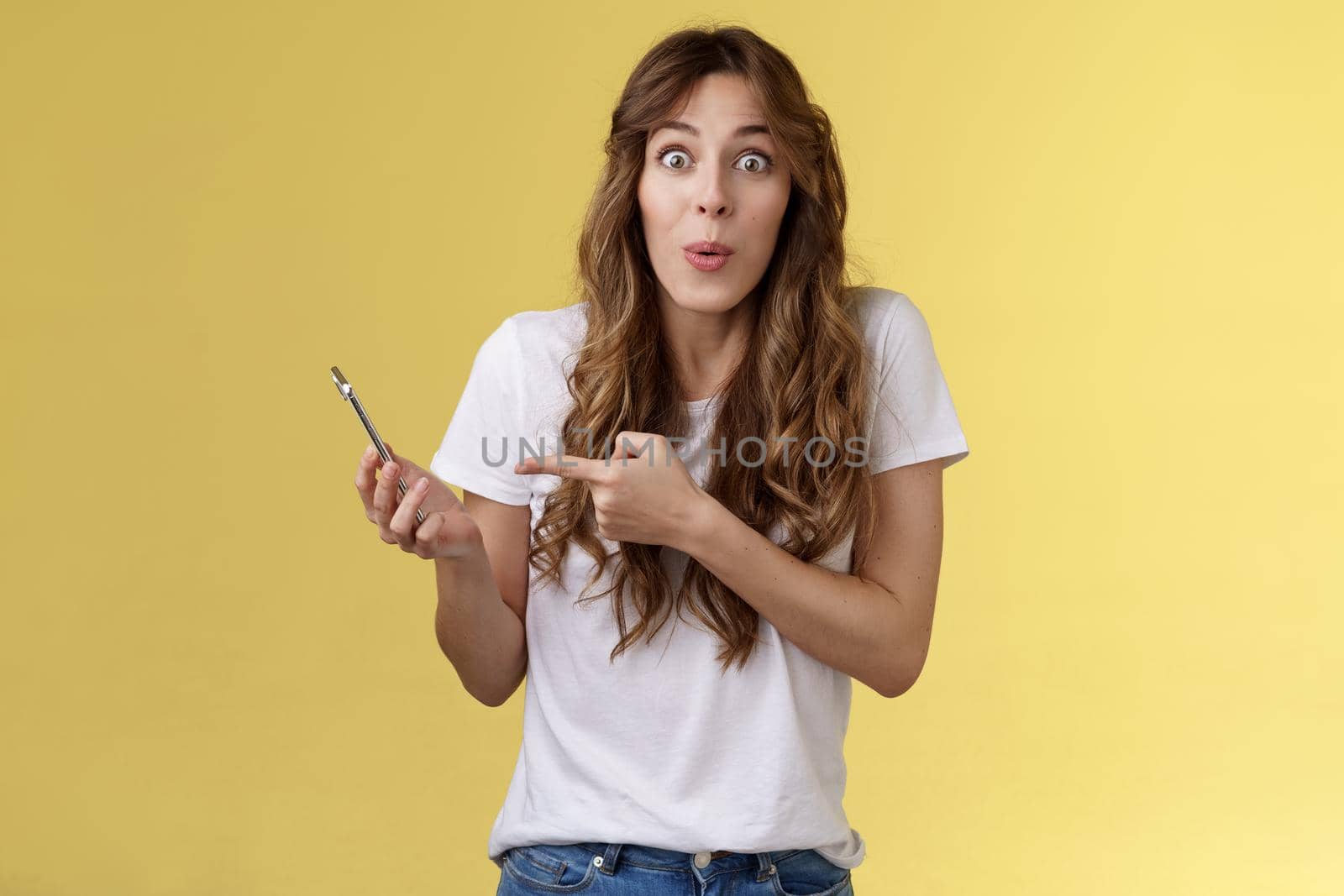 Impressed enthusiastic good-looking girl curly cute hairstyle folding lips amused wow stare camera astonished hold smartphone pointing mobile phone speak about awesome news yellow background. Lifestyle.
