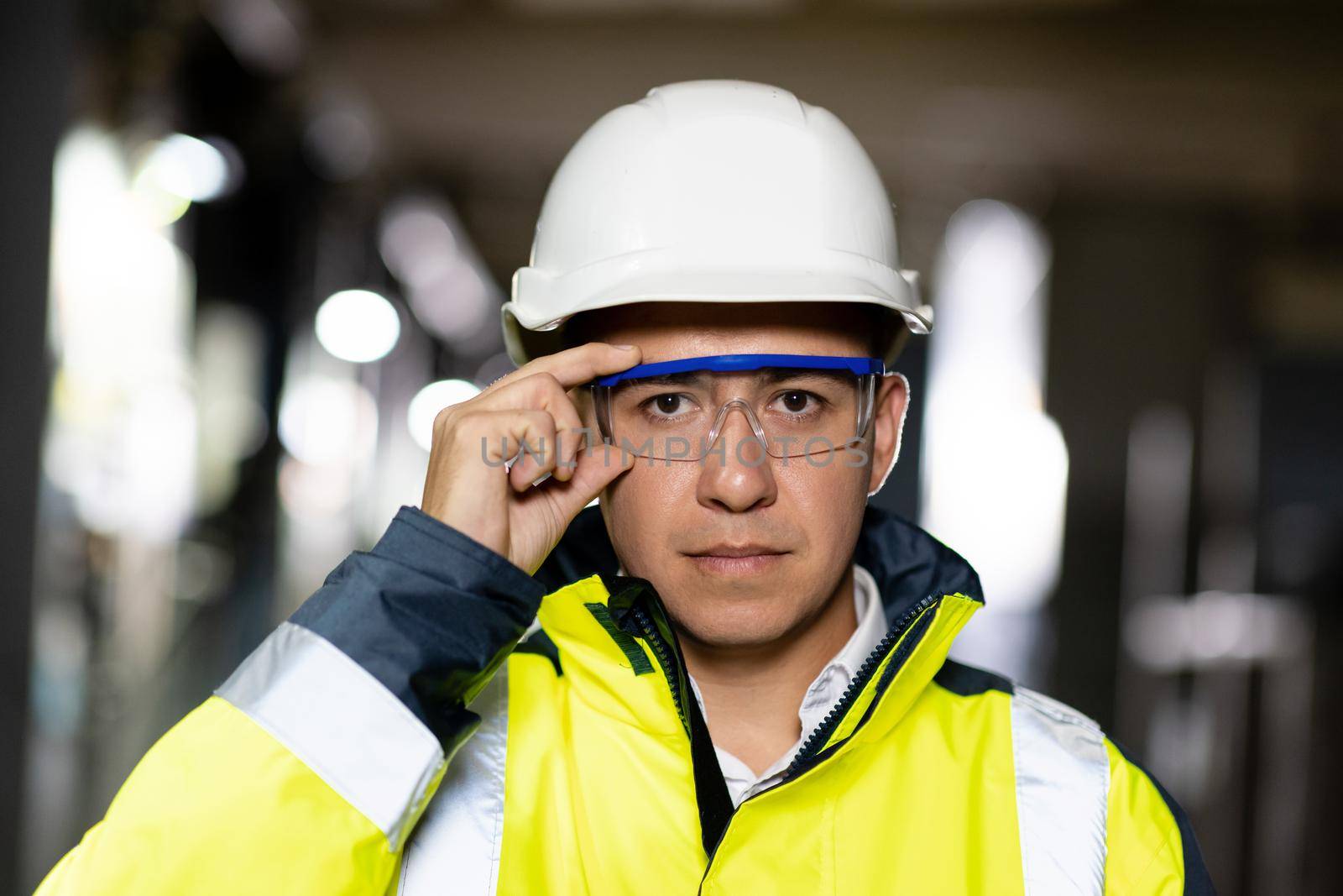 Portrait of employee serious asian man engineer worker wearing safety uniform, goggles and hardhat looking at camera on site factory warehouse background.