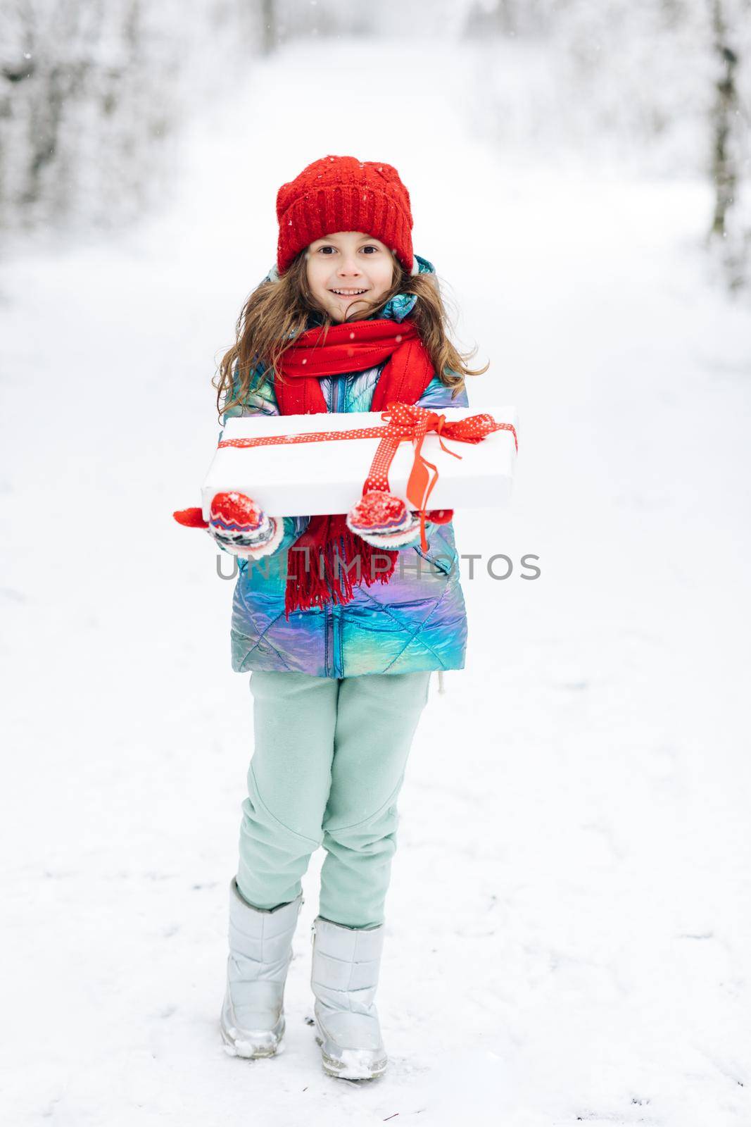 Child in red hat with Christmas presents and gifts in snow. Winter outdoor fun. Kid play in snowy park on Xmas eve. Happy Little caucasian girl smile and holding gift box. by uflypro