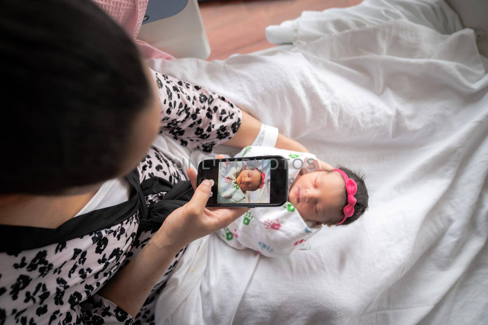 A mother in a pink animal print bathrobe sits on a hospital bed holding her newborn baby girl swaddled in a blanket while taking a photo with her mobile cell phone to share with family and friends. by lapse_life