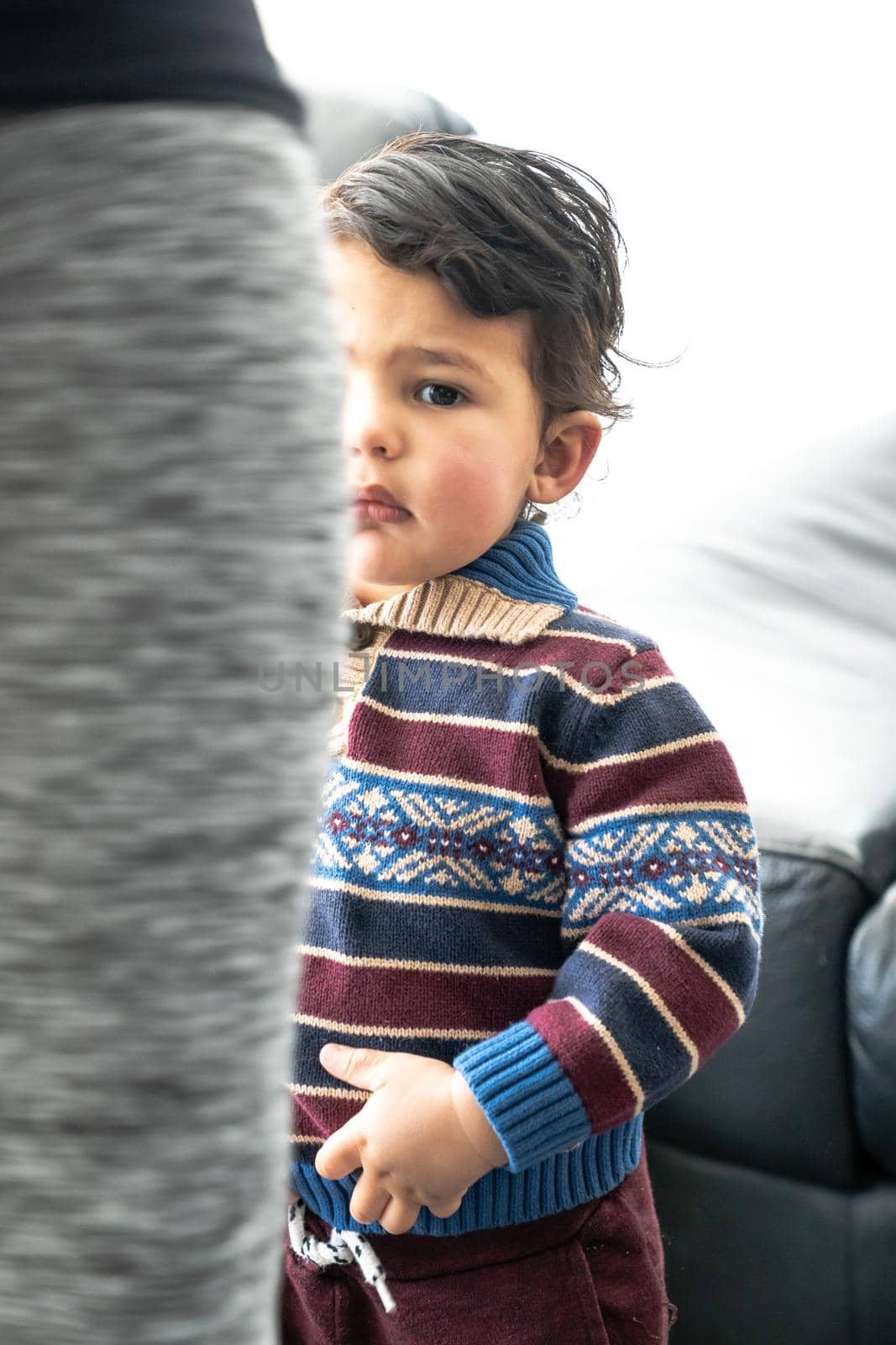 A handsome young baby boy or toddler with long curly brown hair wearing a patterned and striped collared sweater stands by his mother's leg as she combs and fixes the child's hair. by lapse_life