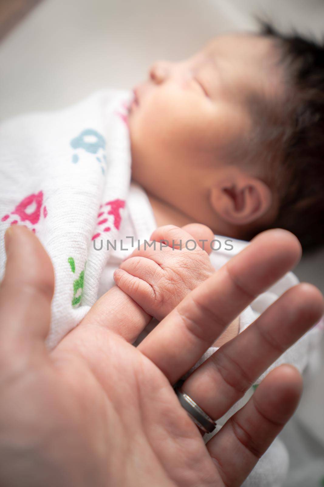 A newborn infant baby girl in a blanket swaddle wraps her tiny hand and fingers around her father's finger as she sleeps peacefully.