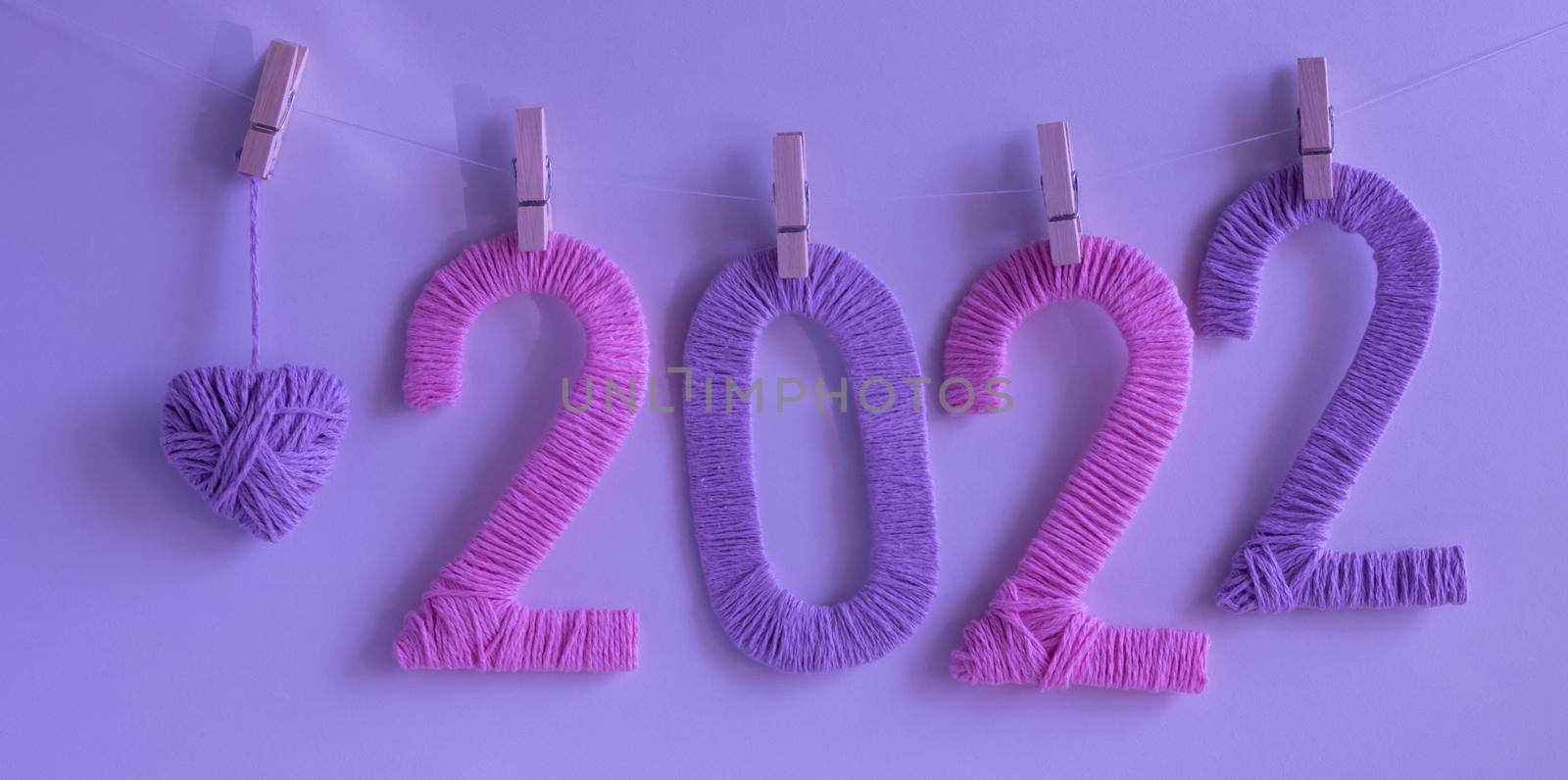 The numbers 2022, made of pink and lilac threads on a neon background, hang on clothespins next to a lilac knitted heart. The concept of the New Year.