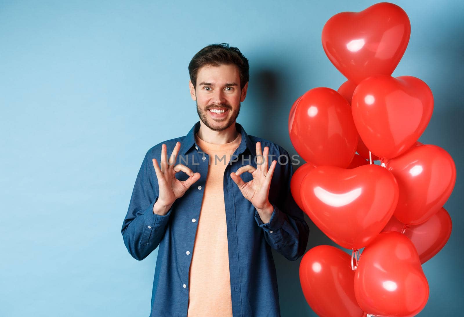 Happy valentines day. Cheerful boyfriend showing okay gestures and praise something good, standing with red hearts balloons for lover, blue background.