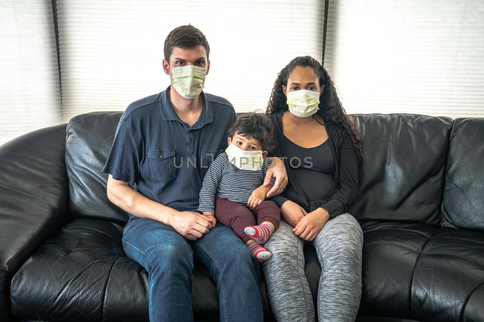 A mixed family sits on a black leather sofa with father and mother holding their infant son all wearing medical face masks in hopes of preventing getting sick from caronavirus or COVID-19. by lapse_life