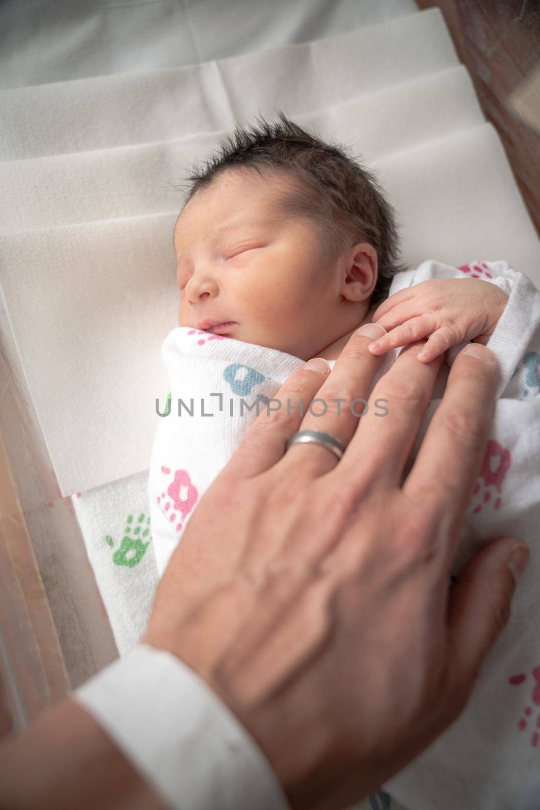 A newborn infant baby girl in a blanket swaddle rests her tiny hand and fingers on her fathers wrinkled hand as she sleeps peacefully.
