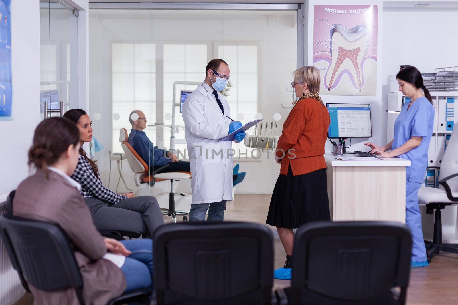 Orthodontist with mask speaking with elderly woman standing in waiting area of stomatological clinic taking notes on clipboard. Nurse typing on computer appointments in modern crowded office.