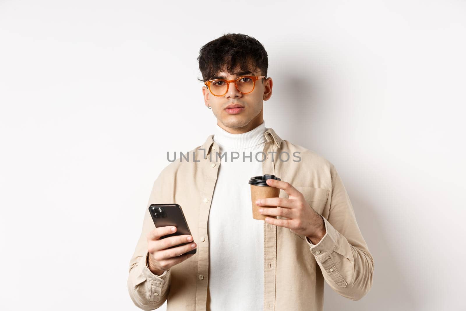 Real people. Stylish hipster guy in glasses holding cup of coffee from cafe takeaway and mobile phone, standing on white background by Benzoix