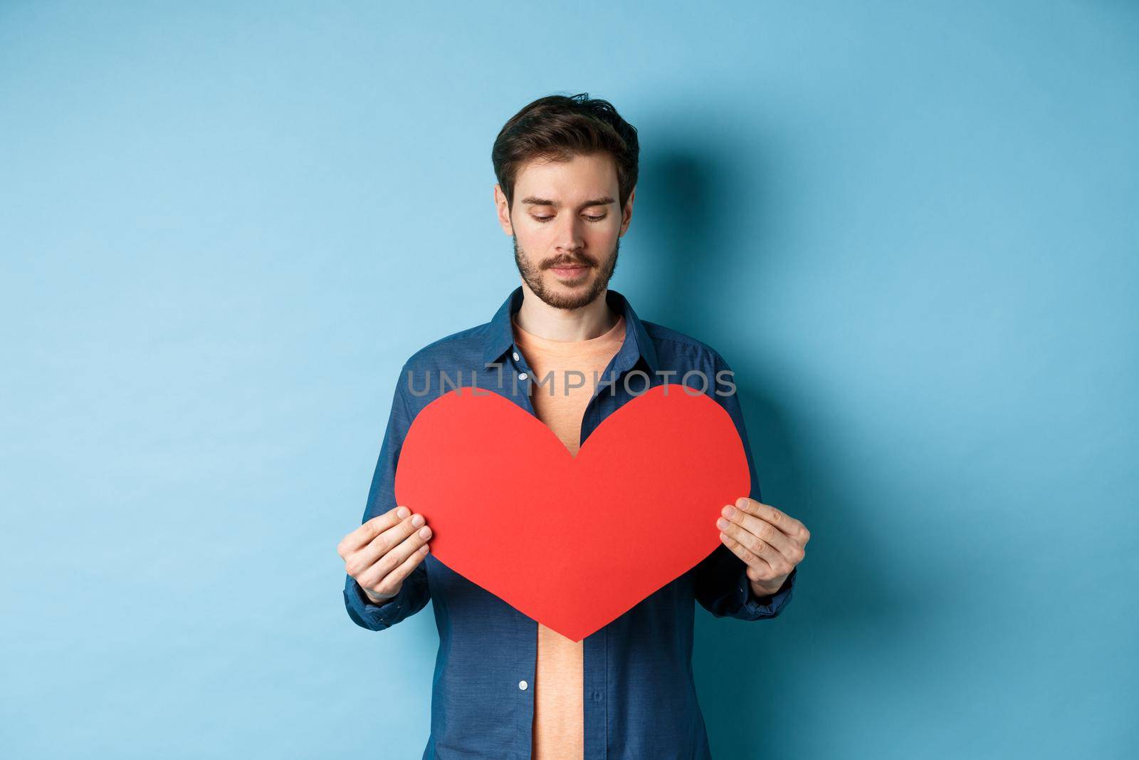 Lonely guy looking sad at valentines red heart with sad face, standing over blue background.