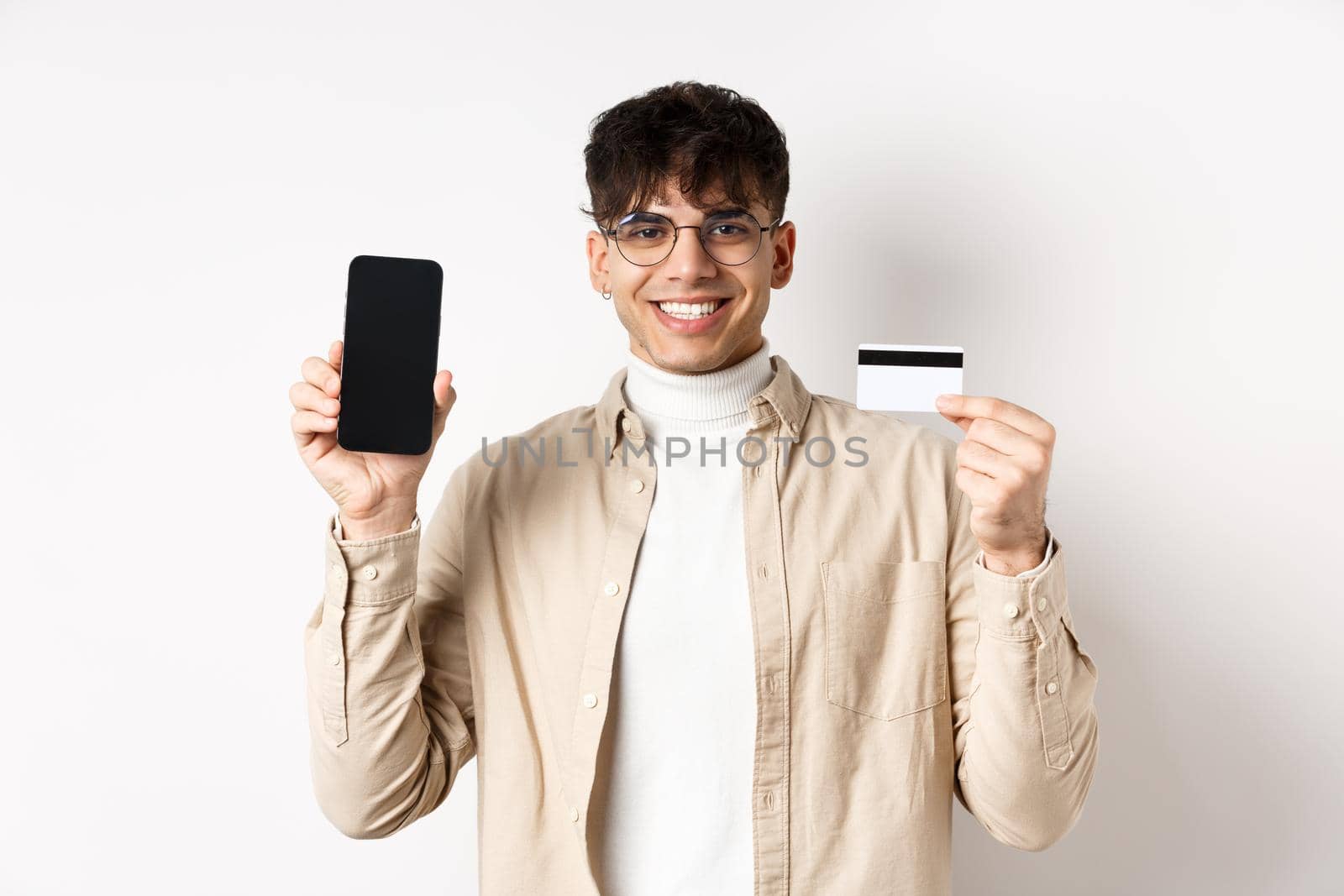 Online shopping. Natural guy in glasses showing empty smartphone screen and plastic credit card, smiling pleased, recommending bank, standing on white background.