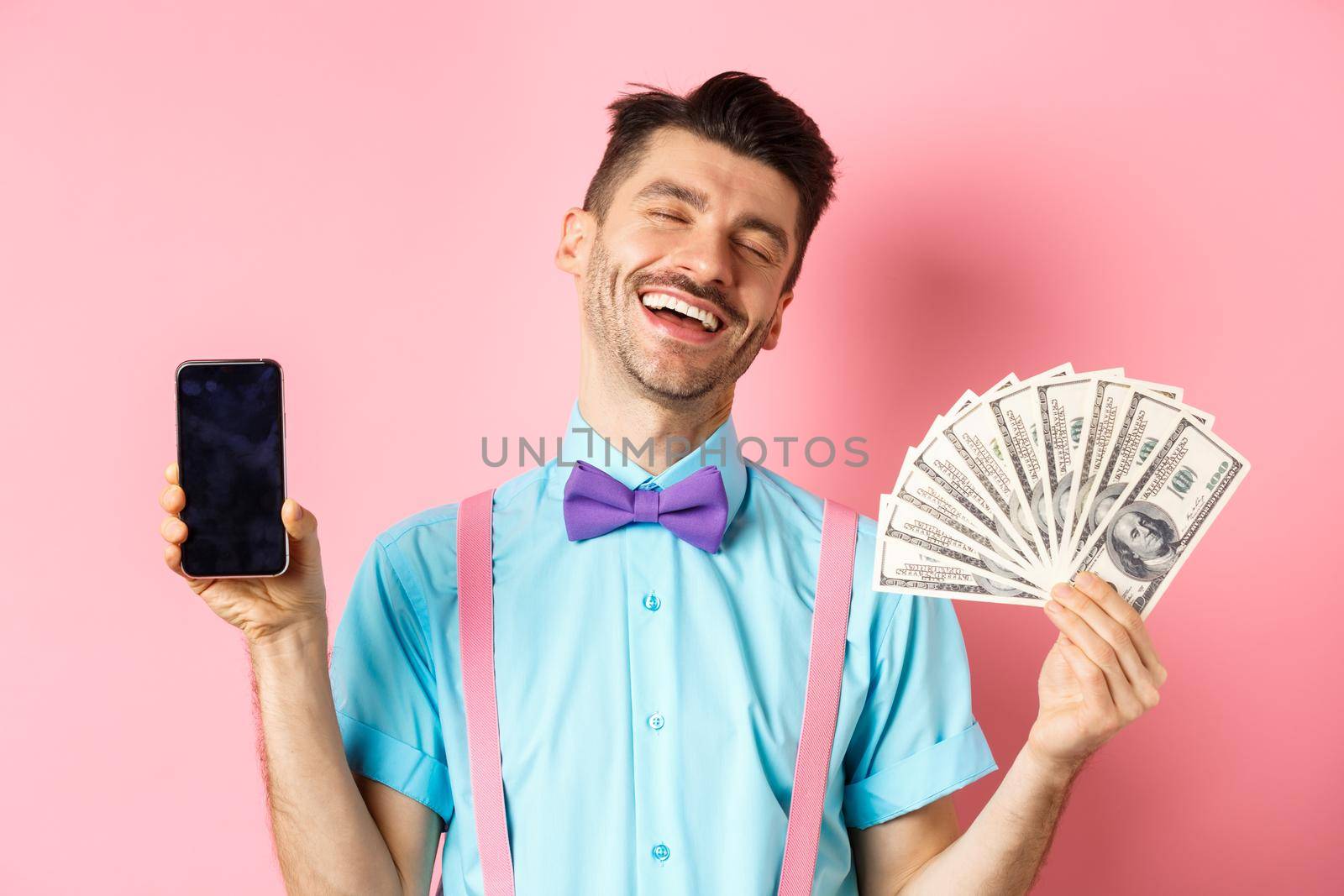 E-commerce and shopping concept. Rich and happy guy laughing, showing money in cash and empty smartphone screen, standing on pink background.