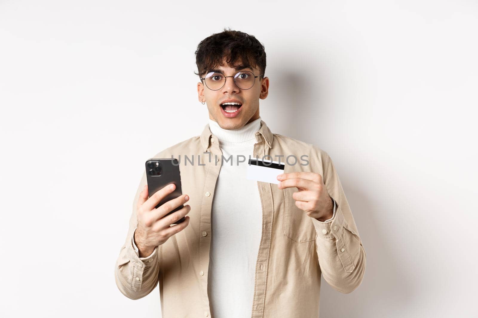 Excited young man shopping online, holding mobile phone and plastic credit card, making purchase in internet, standing on white background by Benzoix