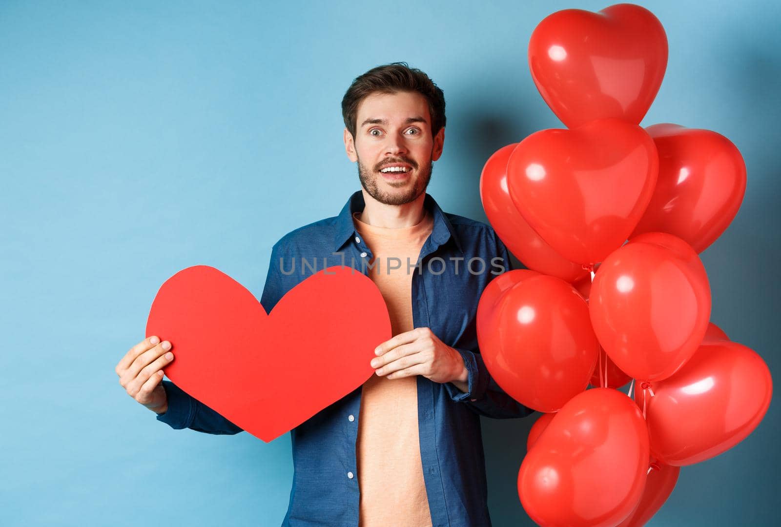 Valentines day concept. Man falling in love, looking startled at girlfriend, showing big red heart and standing near balloon over blue background by Benzoix