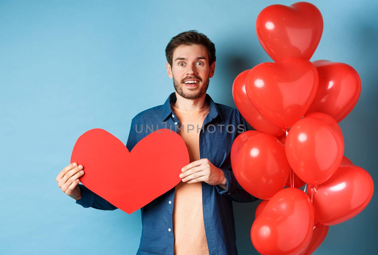 Valentines day concept. Man falling in love, looking startled at girlfriend, showing big red heart and standing near balloon over blue background.