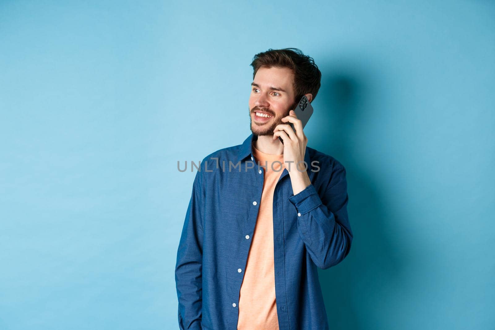 Handsome caucasian man talking on mobile phone, looking aside at empty space with carefree smile, standing on blue background by Benzoix