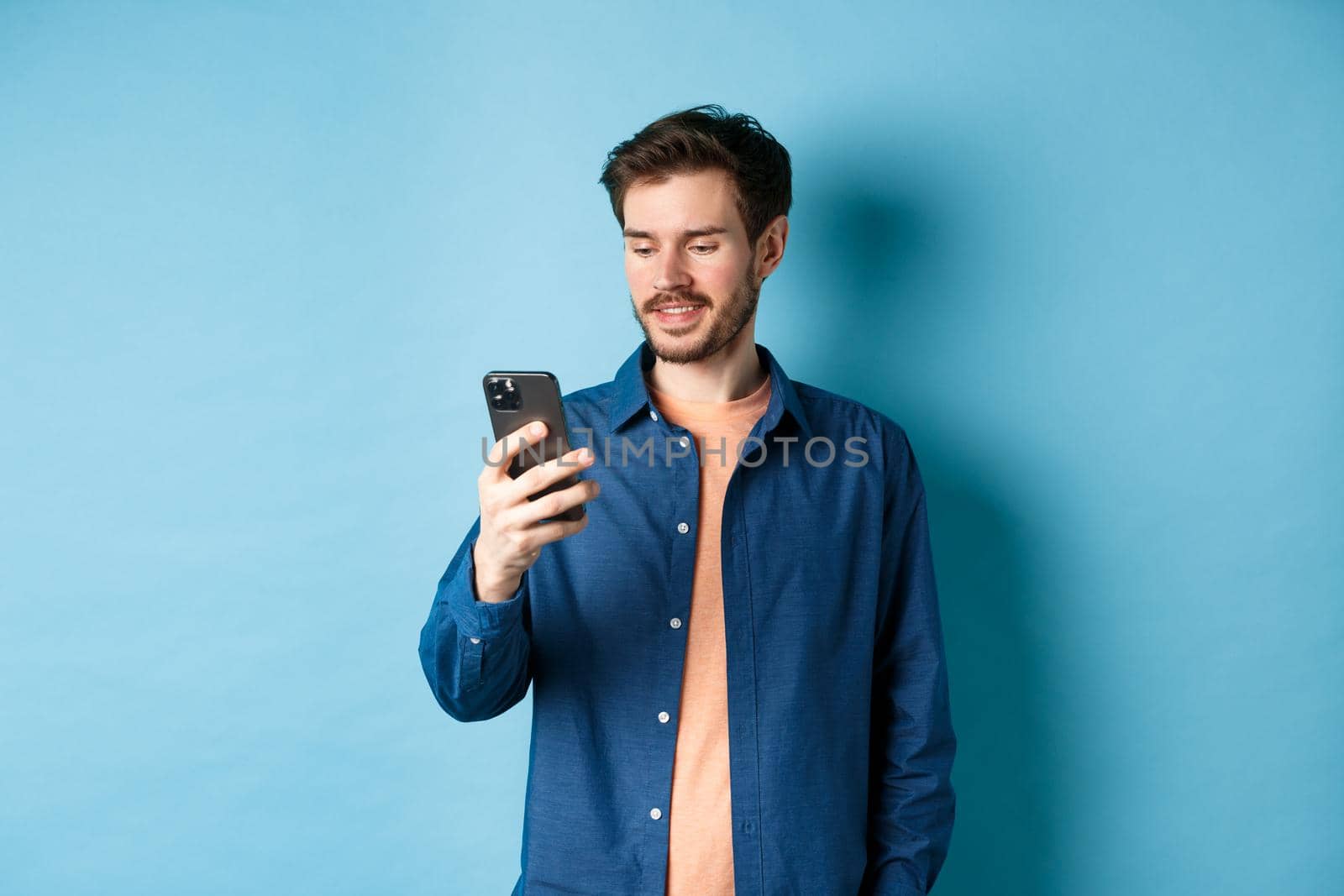 Handsome modern guy using mobile phone, reading smartphone screen and smiling, networking on blue background by Benzoix