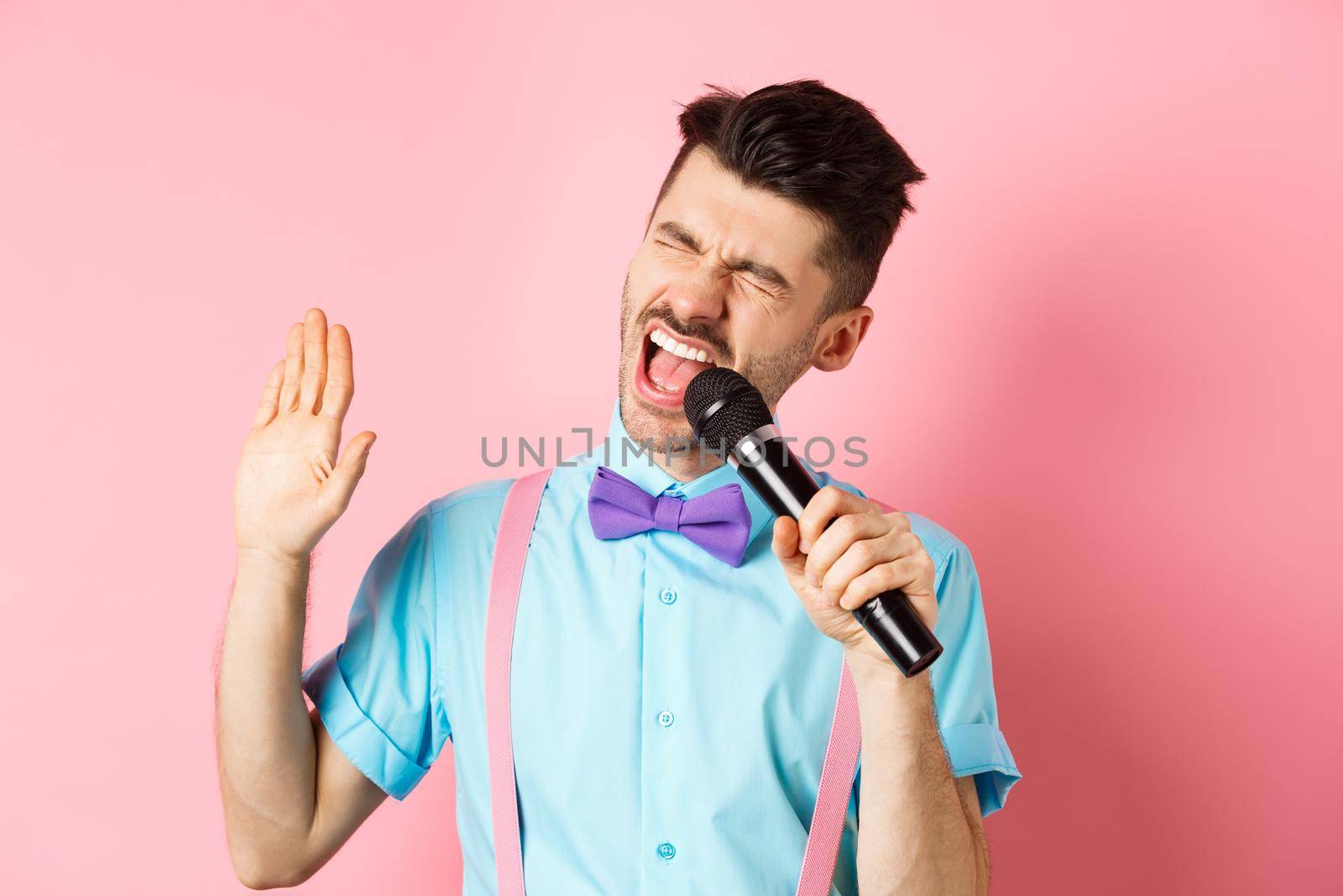 Party and festive events concept. Funny guy performs a song, singing karaoke in microphone with carefree face, standing in bow-tie and suspenders on pink background.
