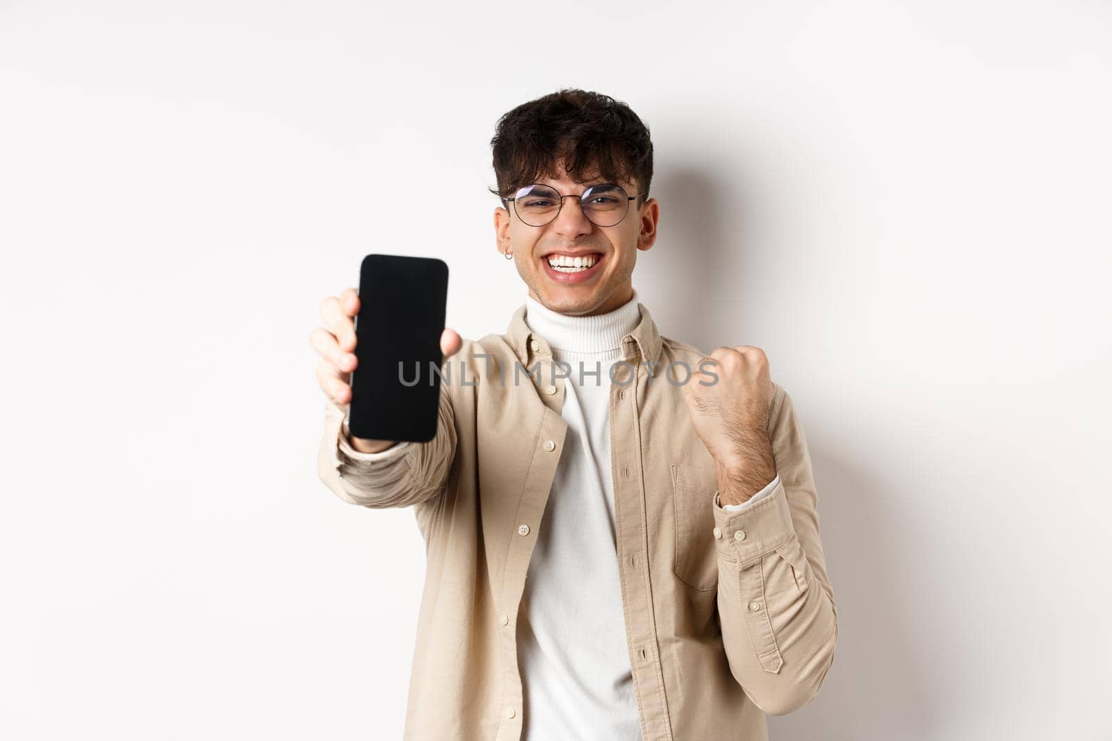 Lucky young man winning prize online, showing smartphone screen and rejoicing of good news, standing happy on white background by Benzoix