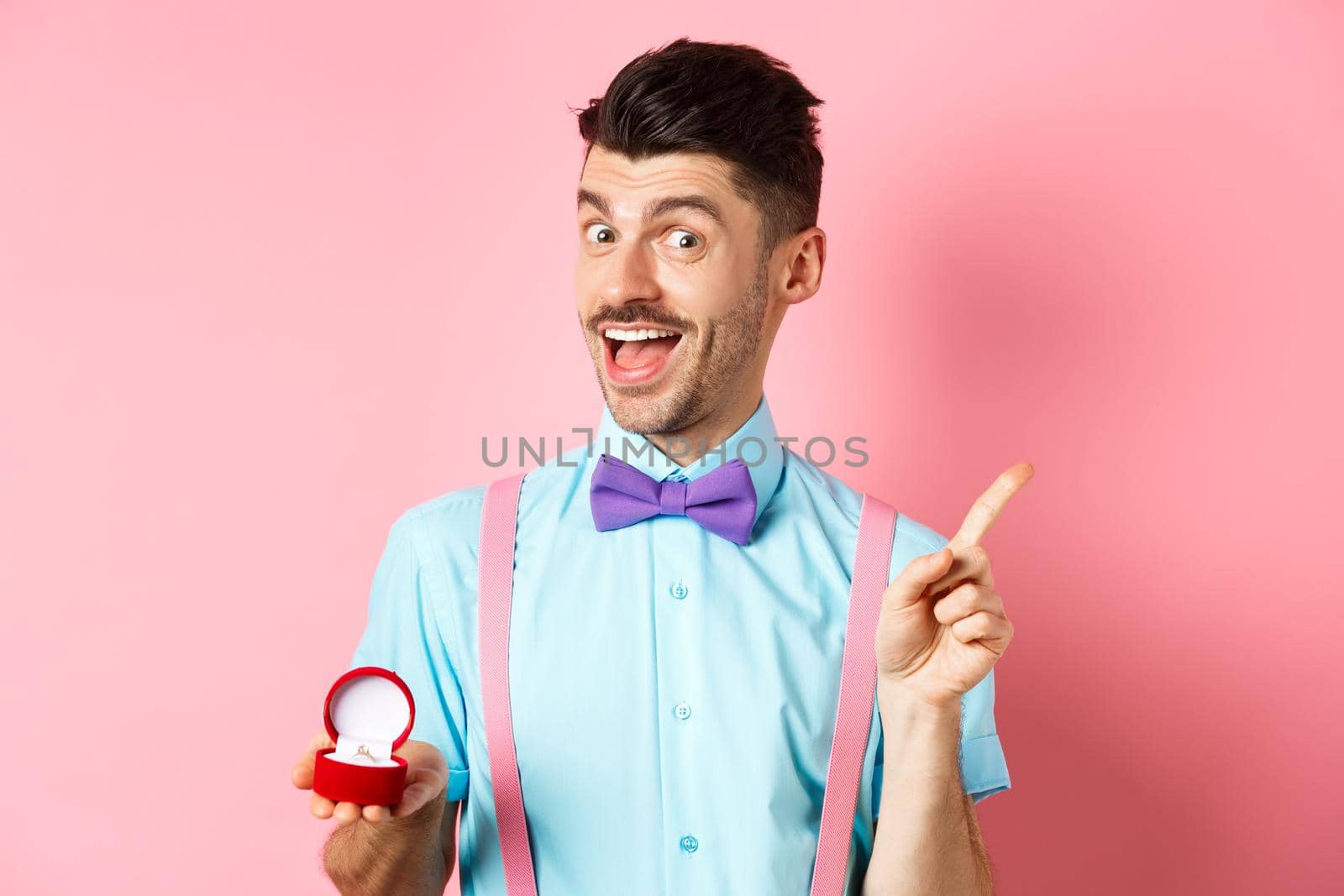 Valentines day. Funny guy with moustache and bow-tie, showing engagement ring and pointing finger at upper right corner, standing on pink background.