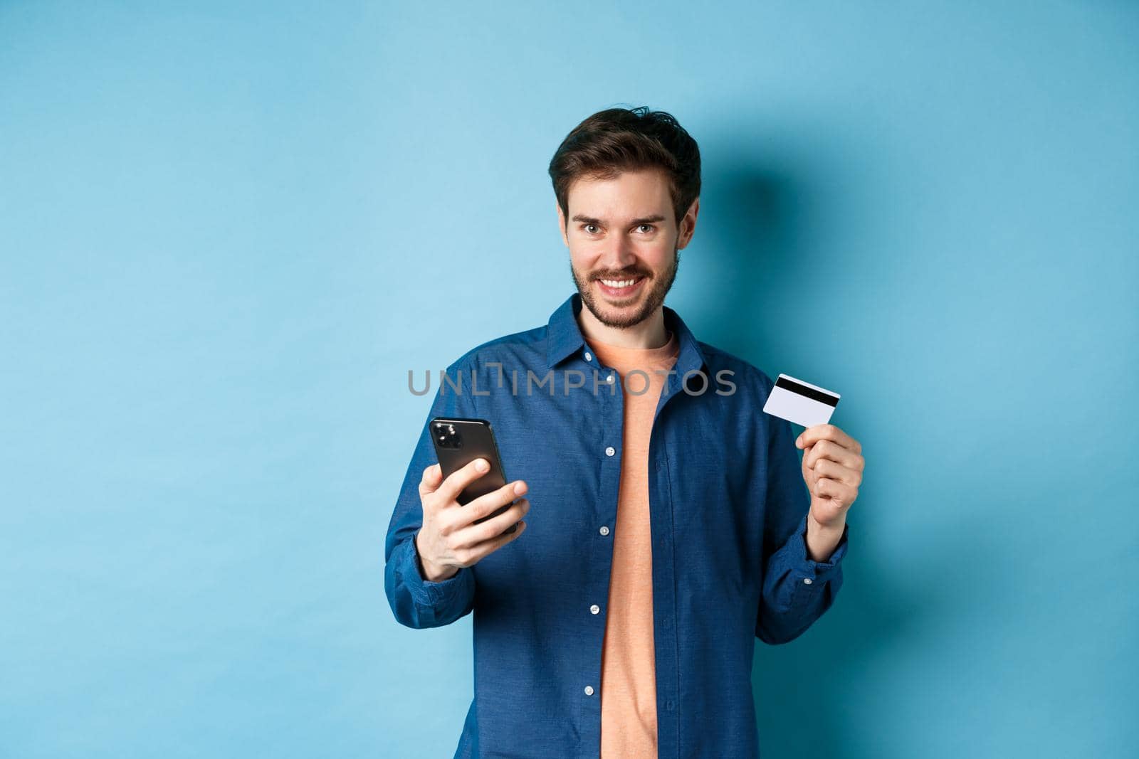 E-commerce concept. Handsome young man shopping online, holding smartphone and plastic credit card, smiling at camera, standing on blue background.