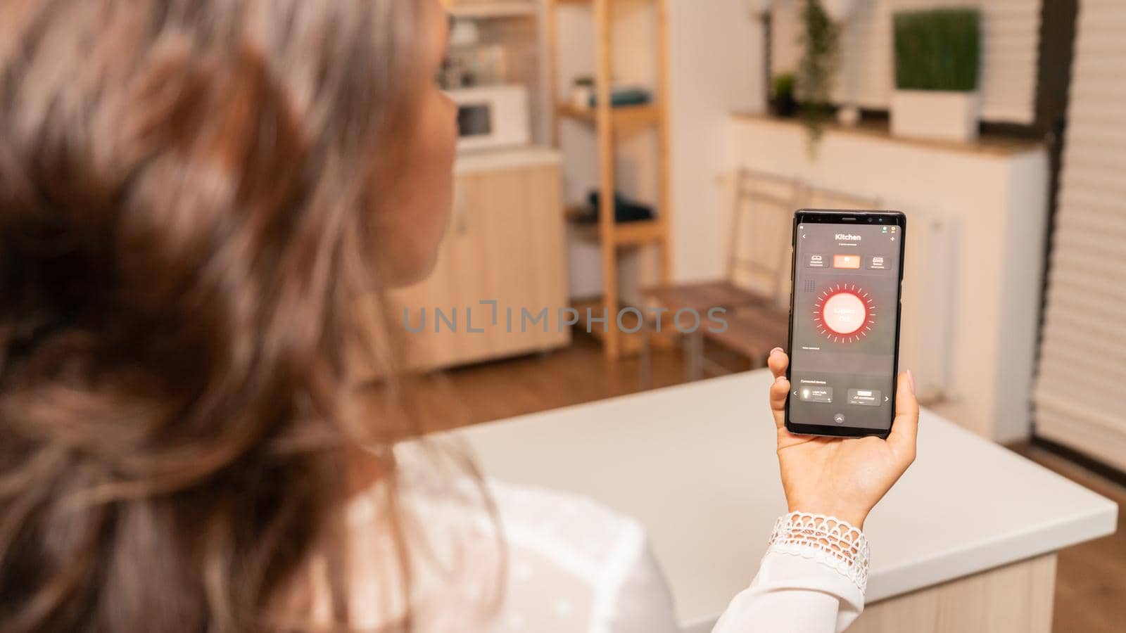Woman in kitchen using voice controll on phone to change lighting. Person in apartment holding telephone with touchscreen and app for lights.