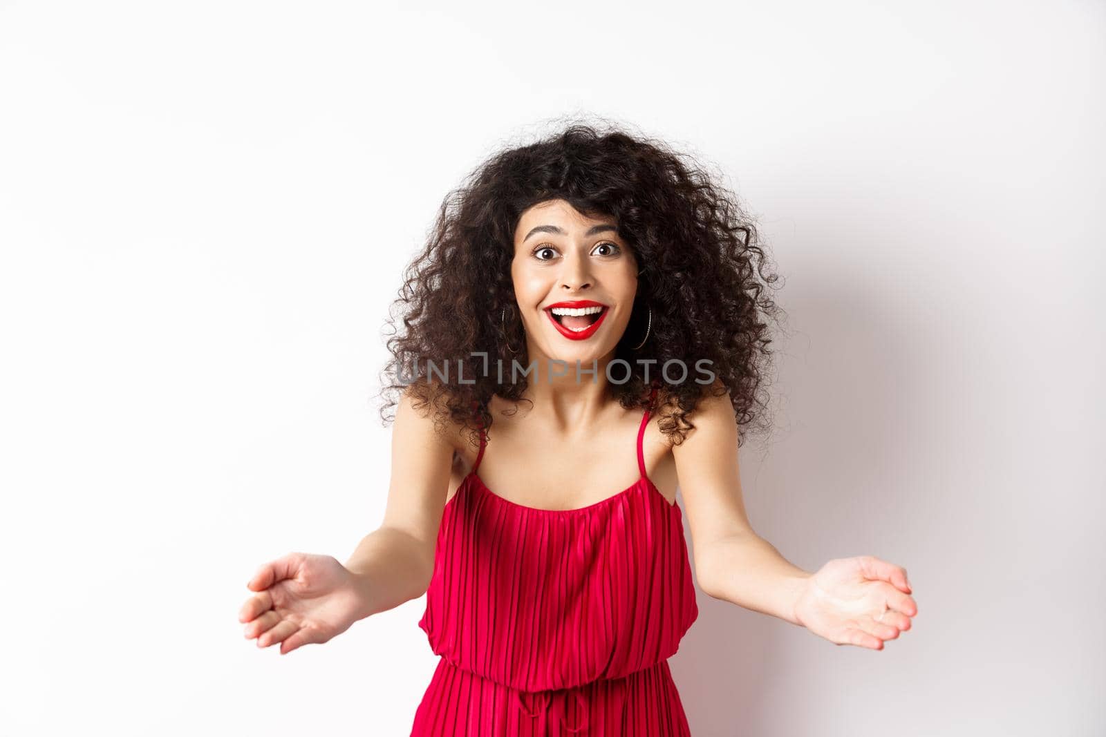 Portrait of happy caucasian woman in red dress and makeup, stretch out hands to beckon someone, inviting for hug, receiving surprise gift, standing on white background.