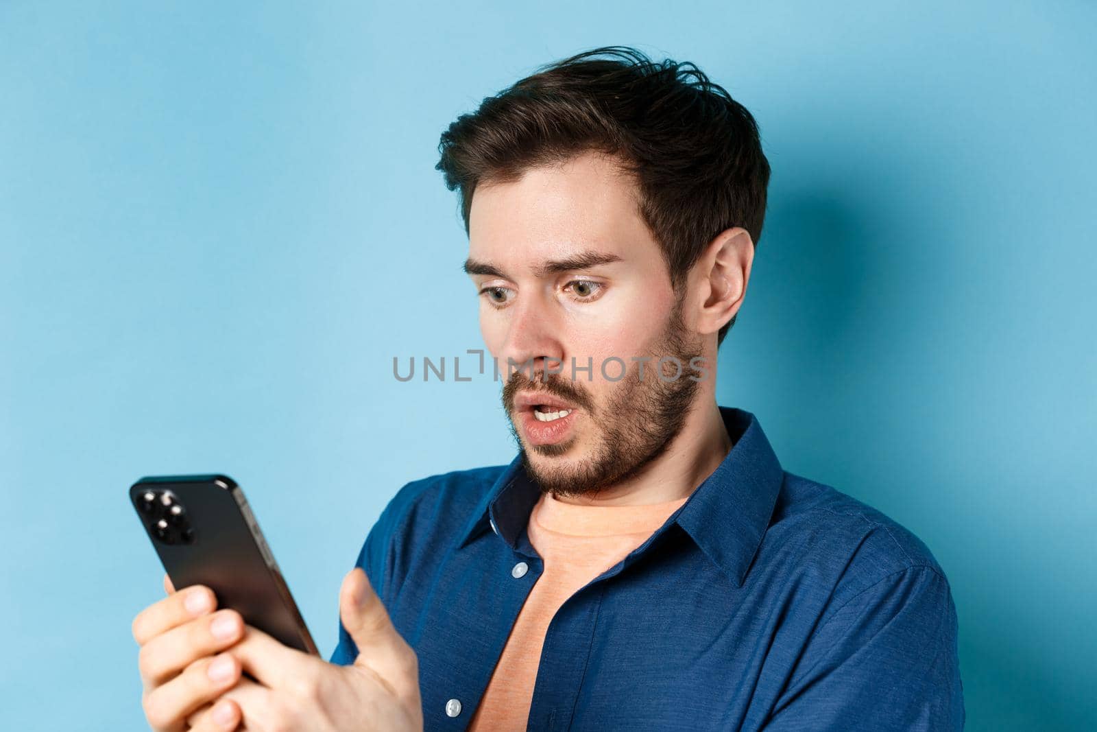 Close-up of man looking shocked and reading message on smartphone, gasping and stare startled at screen, standing on blue background.