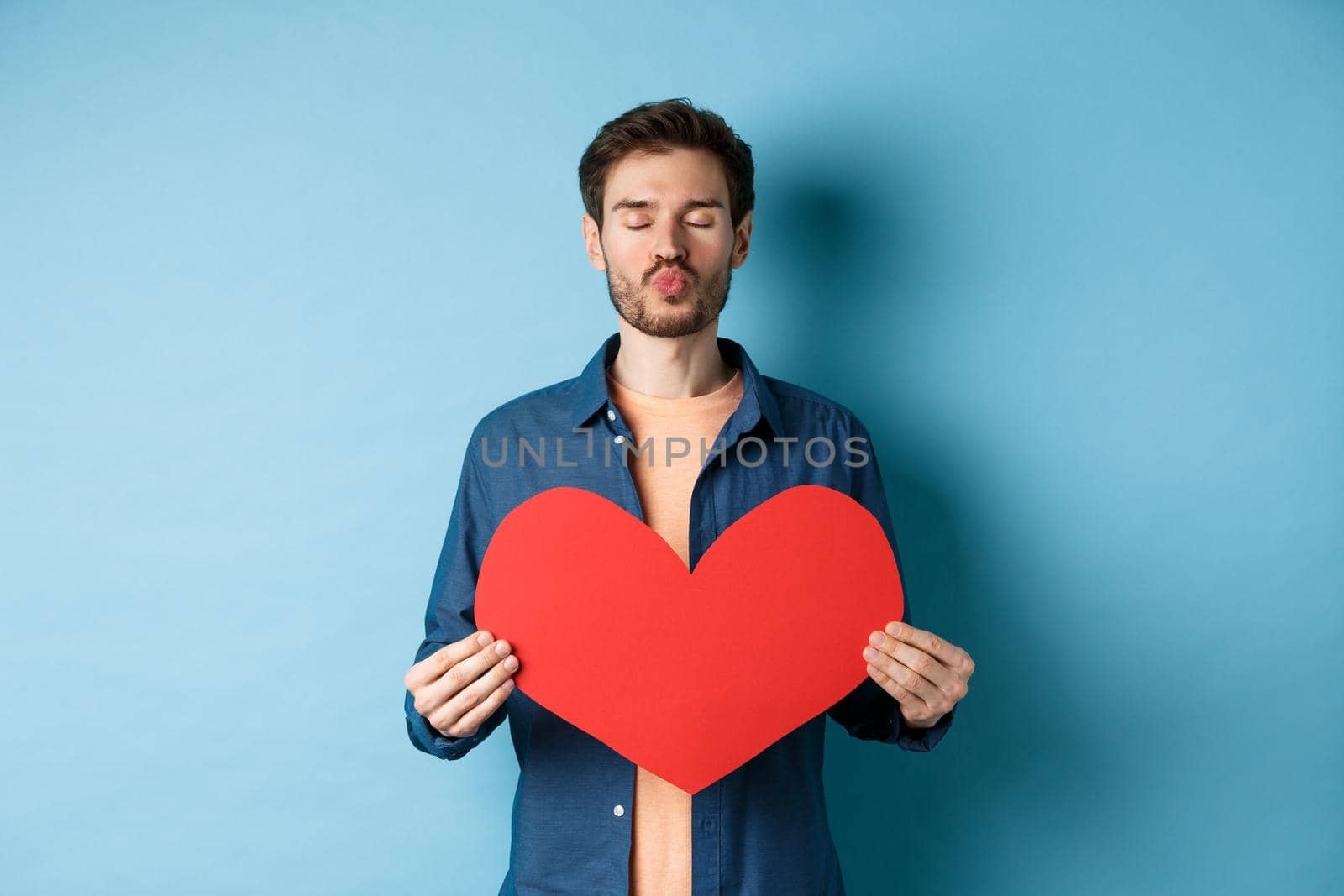 Romantic boyfriend with valentines red heart close eyes, pucker lips and waiting for kiss on lovers day, standing against blue background.