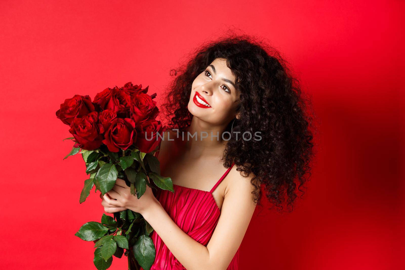 Happy beautiful woman in dress, holding flowers and smiling romantic, looking aside at logo, standing against red background.