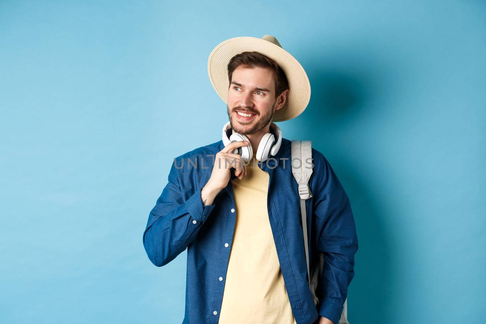 Handsome tourist with backpack and straw hat looking aside, smiling and touching headphones, standing on blue background.