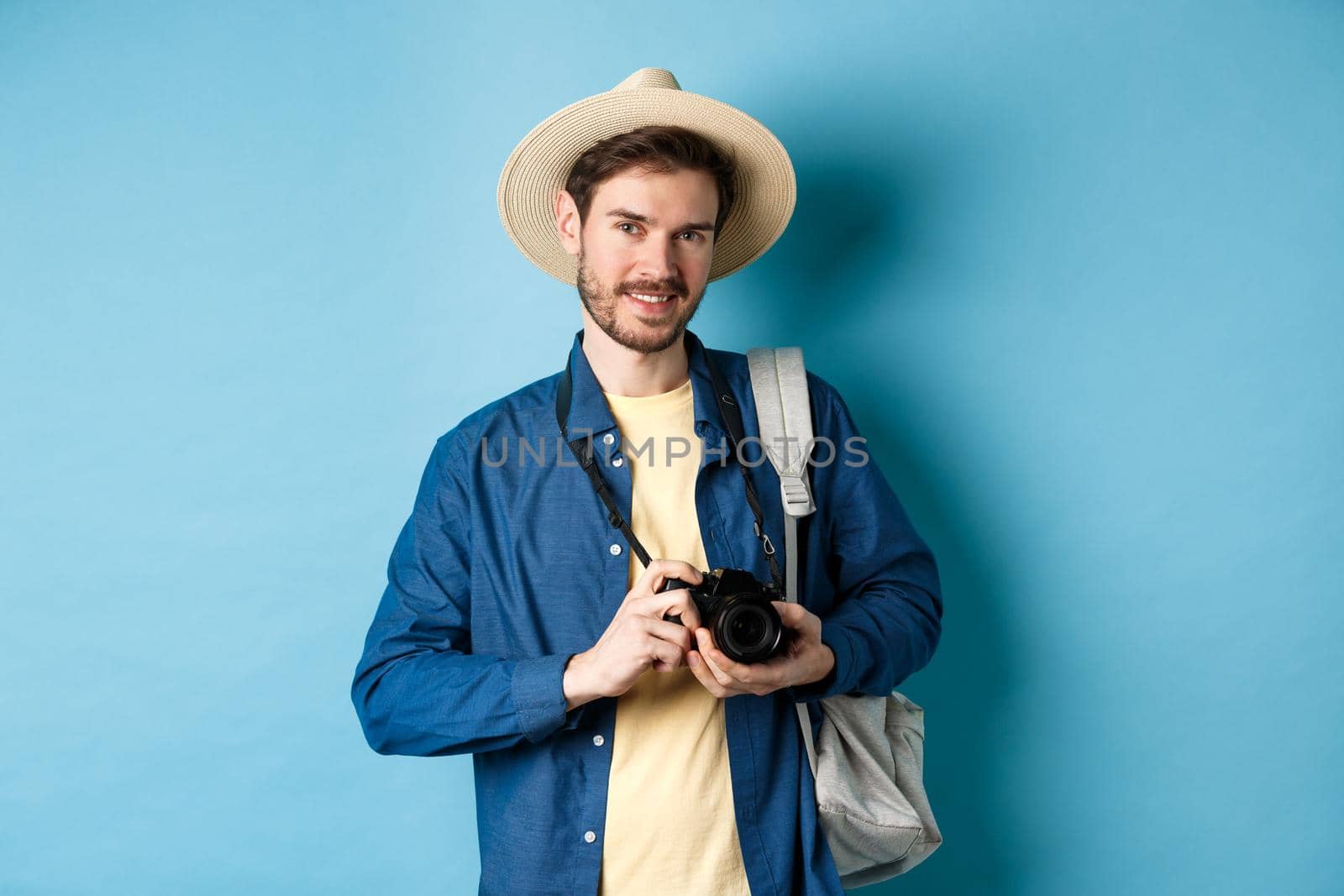 Handsome guy going on summer vacation, backpacking on holiday. Tourist with straw hat and camera smiling happy, standing on blue background.