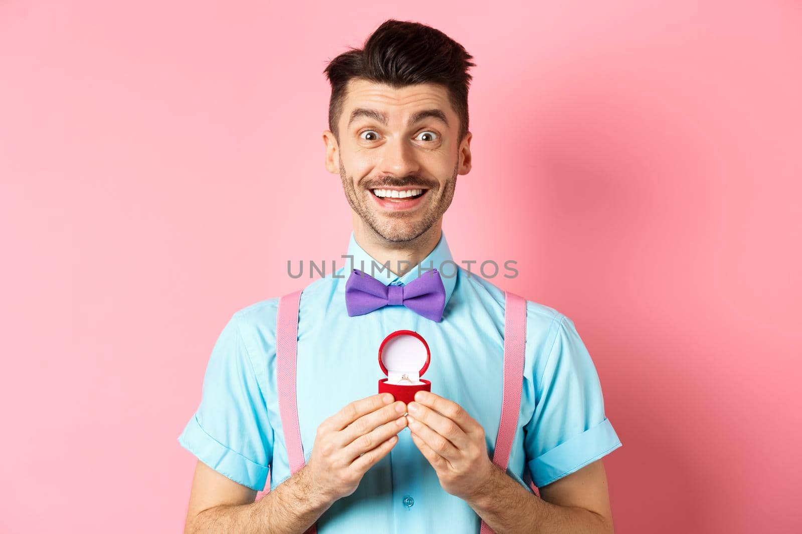 Valentines day. Romantic man in bow-tie showing engagement ring and smiling, asking to marry him, making proposal to lover on pink background by Benzoix