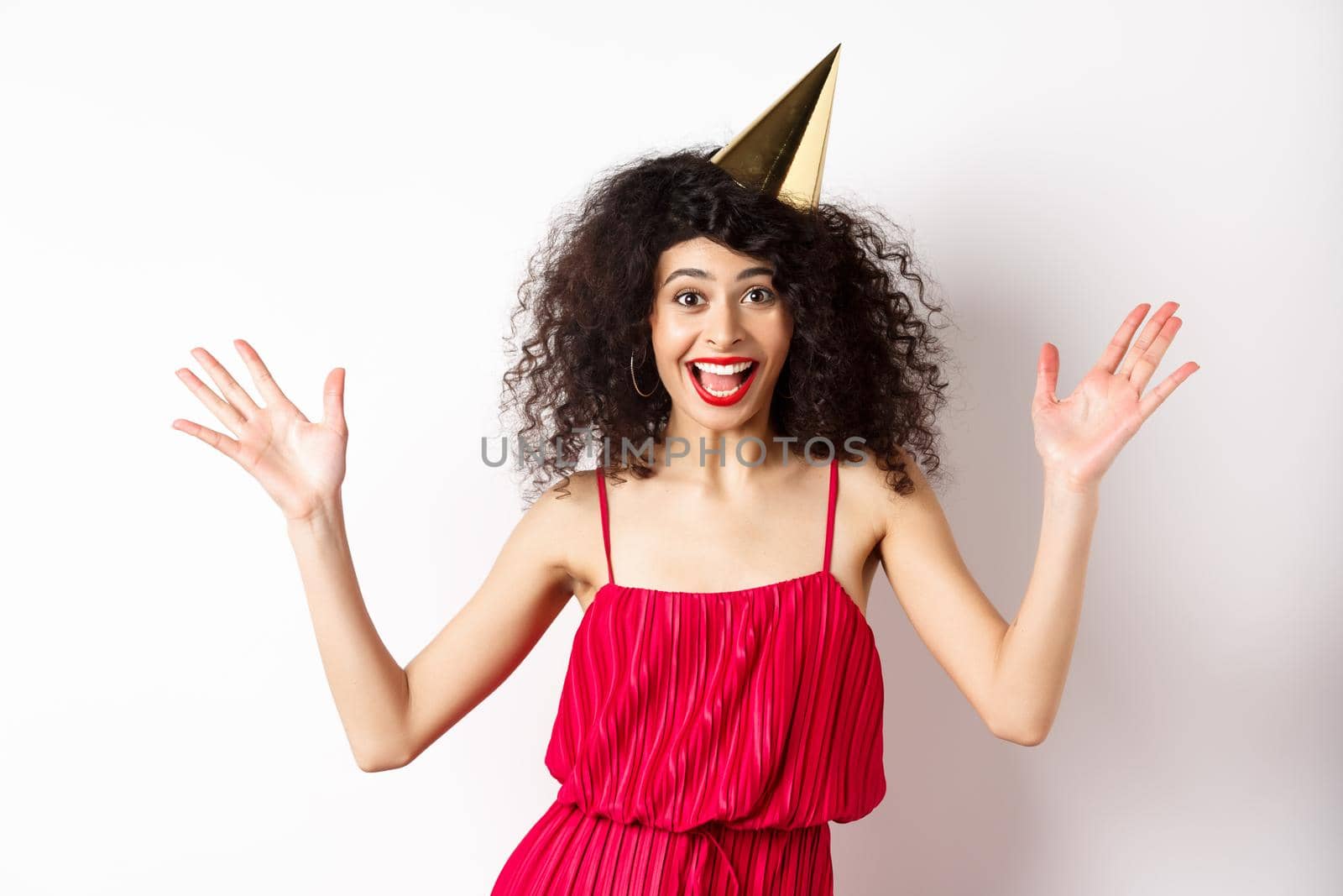 Cheerful young woman in red dress, celebrating birthday, wearing party hat and smiling, screaming of joy, standing on white background by Benzoix