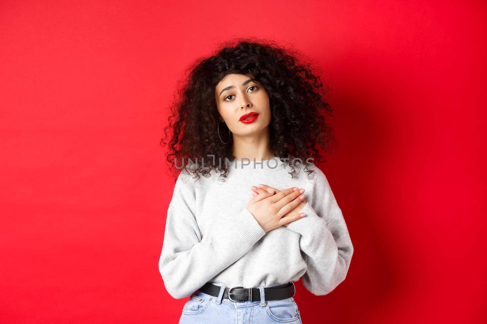 Beautiful woman with curly hair feeling touched and thankful, holding hands on heart and looking with affection at camera, red background.