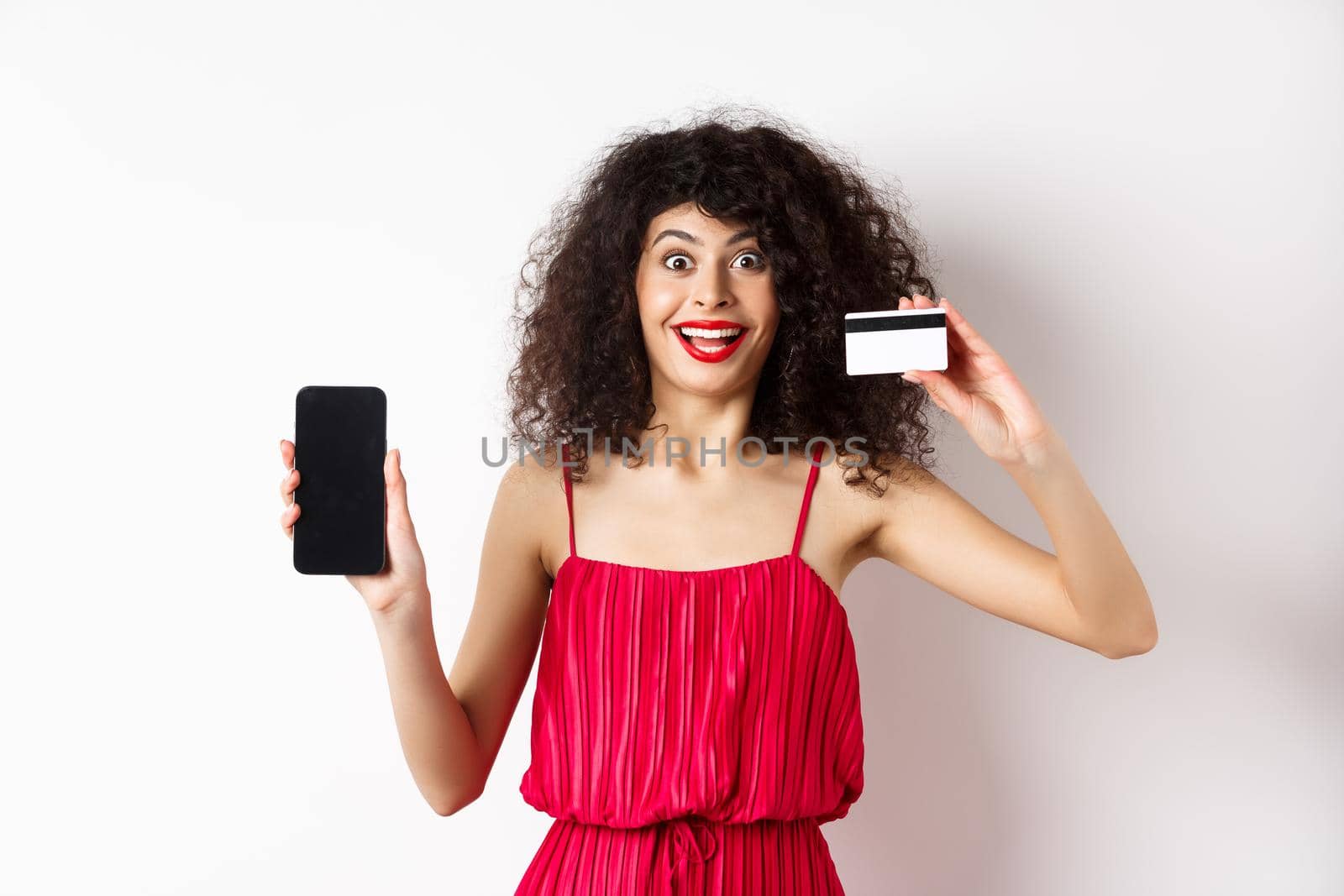 Online shopping concept. Excited curly-haired woman in red dress showing empty smartphone screen and plastic credit card, smiling happy at camera, standing on white background by Benzoix