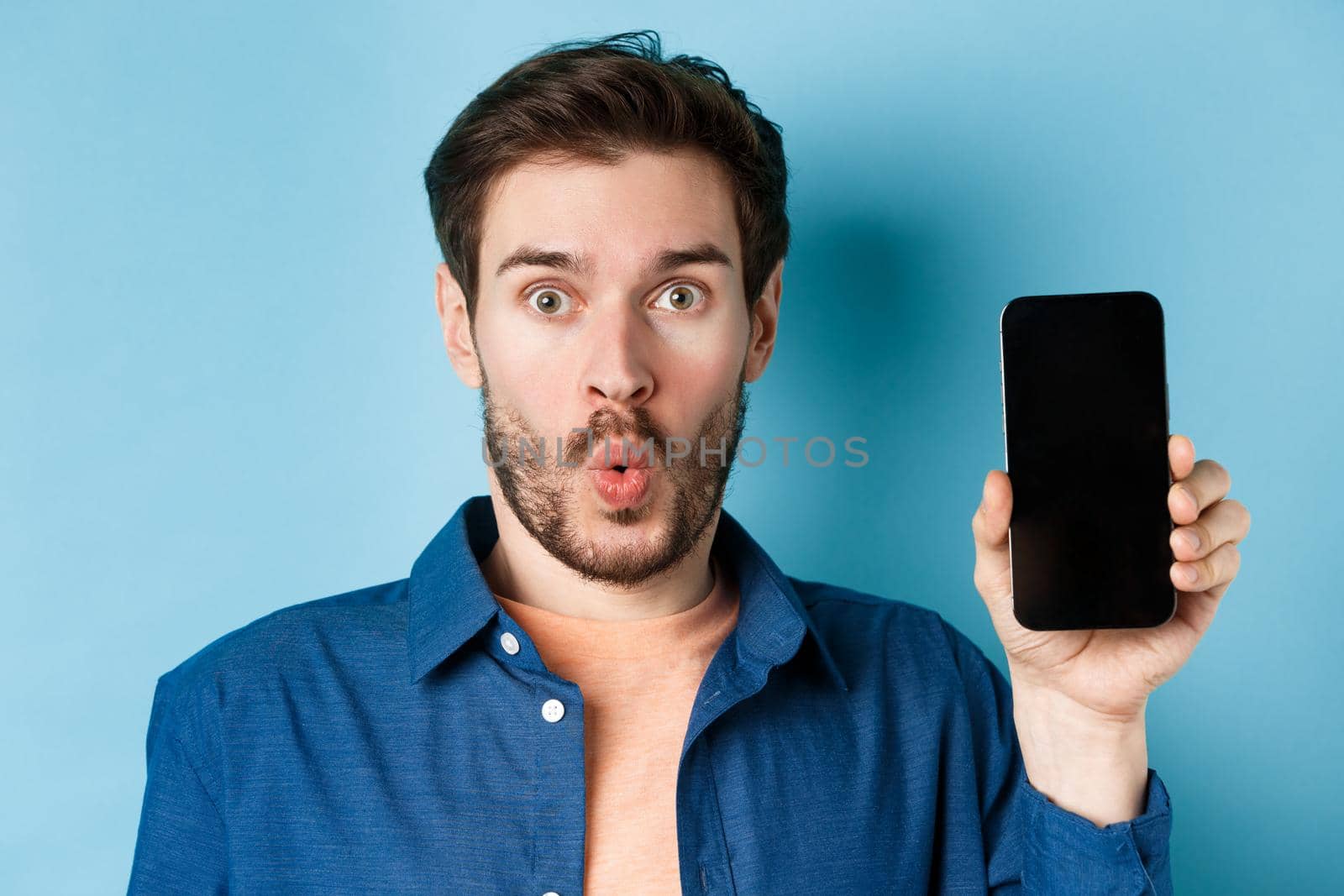 Close up portrait of impressed man say wow, showing empty mobile phone screen, standing on blue background.