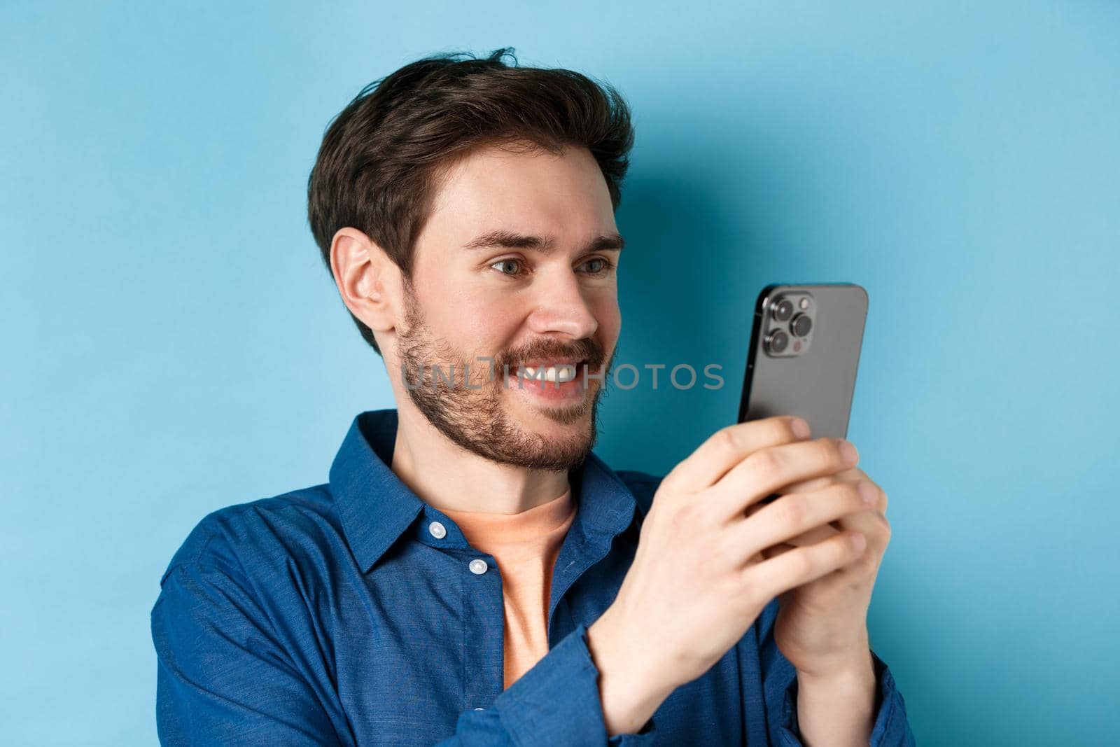 Image of handsome caucasian man reading mobile screen, taking pictures on smartphone and smiling, standing on blue background.