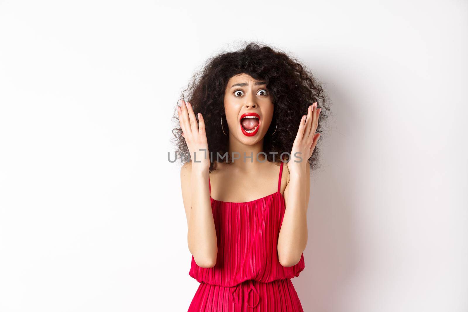 Woman scream in panic, wearing red dress and shouting at camera with anxious face, standing over white background. Copy space