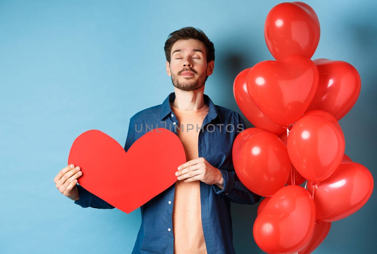 Valentines day concept. Man dreaming of true love, holding red heart cutout and standing near romantic balloons, blue background by Benzoix
