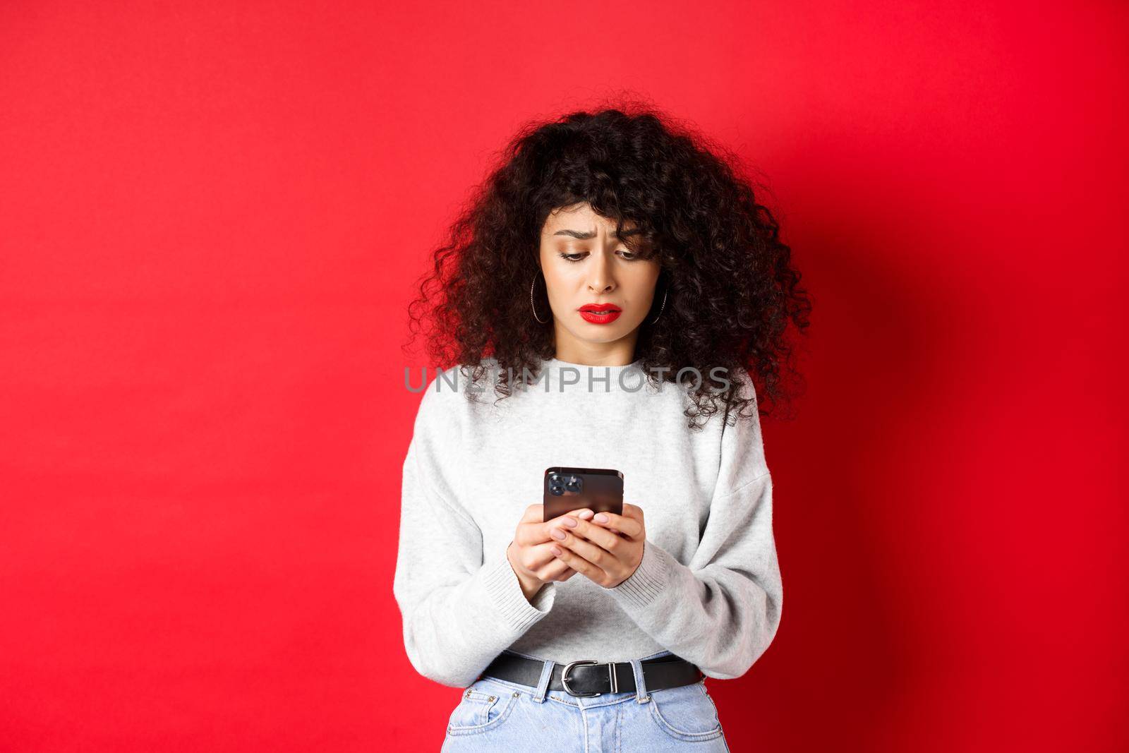 Sad and worried woman reading message on smartphone, receive bad news on social media, standing against red background by Benzoix