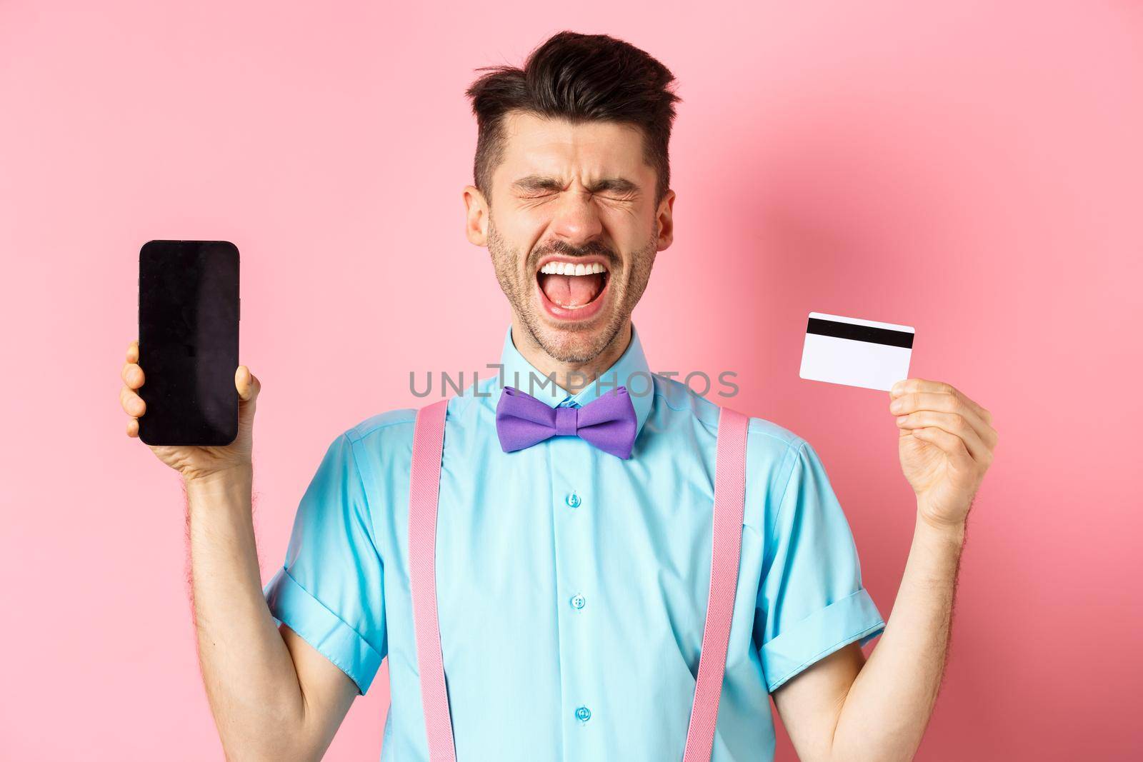 E-commerce and shopping concept. Crying man showing empty smartphone screen and plastic credit card, feeling frustrated, standing on pink background.