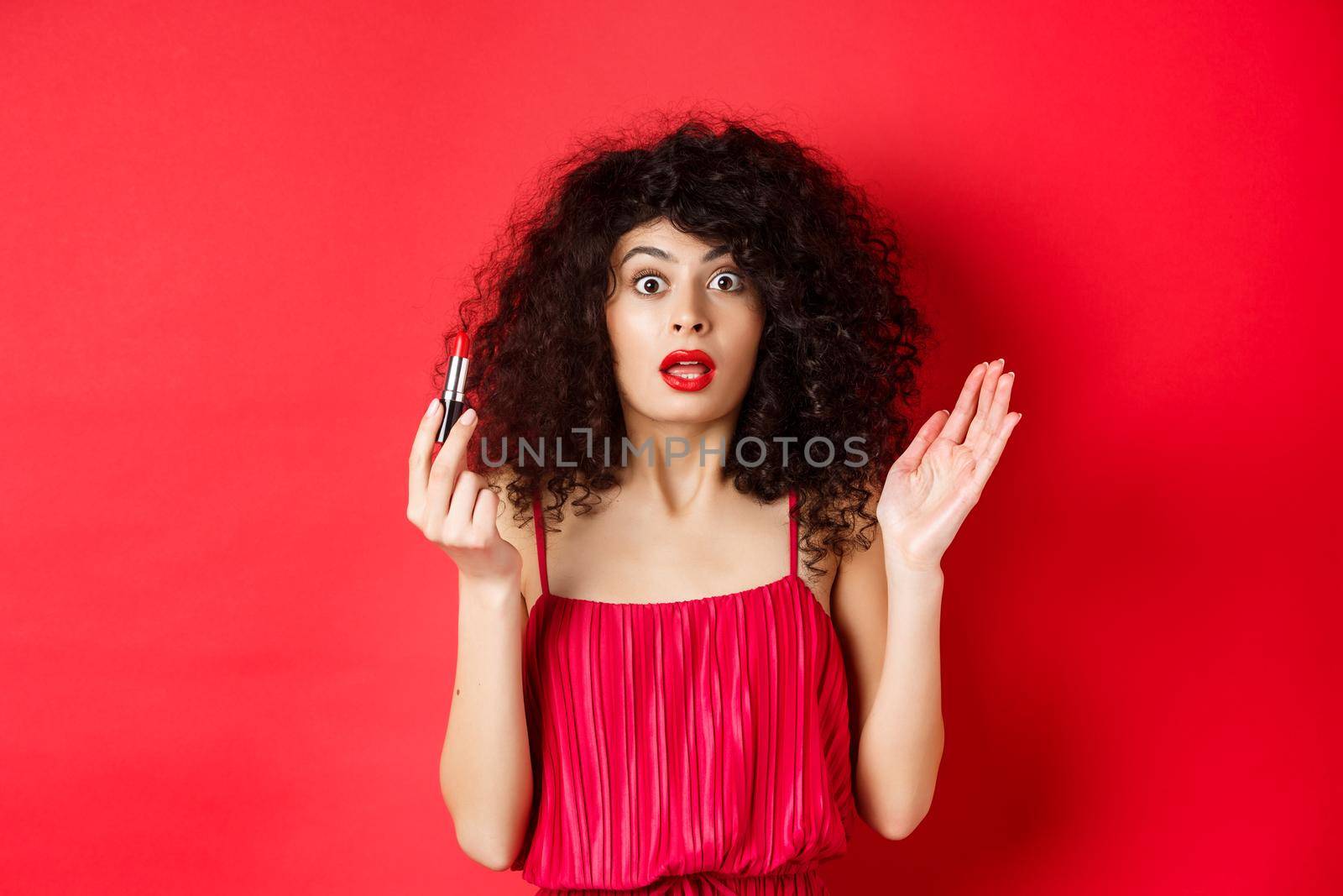 Beauty and makeup concept. Excited woman with curly hair, gasping as looking at camera, holding red lipstick, standing in dress on white background.