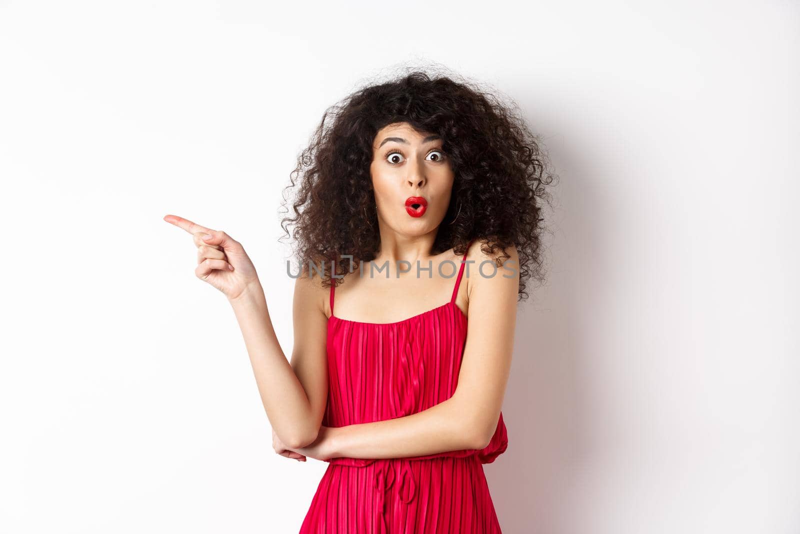 Surprised elegant woman in red dress, saying wow and pointing finger left, showing interesting promo, standing over white background.