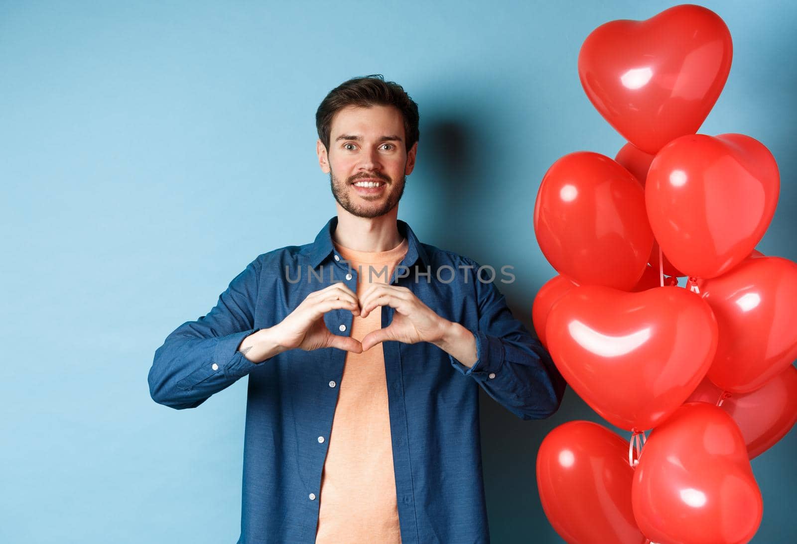Happy valentines day. Boyfriend in love showing heart gesture to lover and smiling, standing near red balloons on blue background by Benzoix