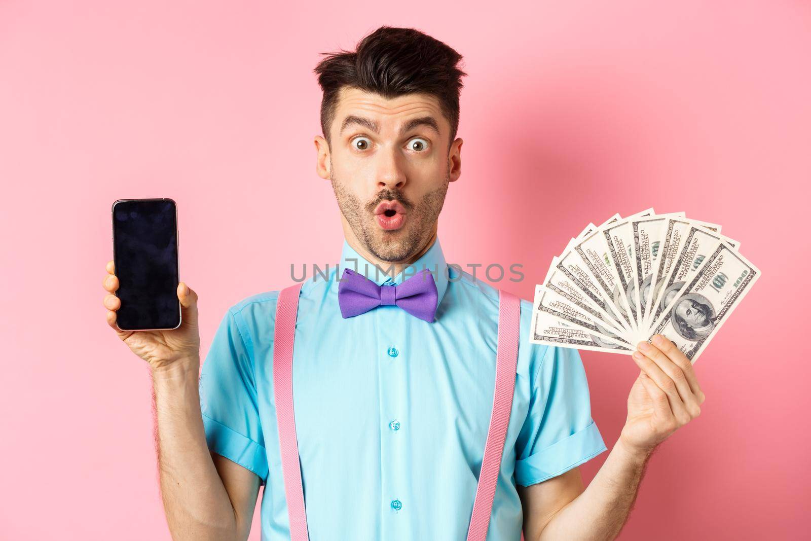 E-commerce and shopping concept. Surprised man showing blank smartphone screen and money, saying wow with amazed face, checking out online offer, pink background by Benzoix