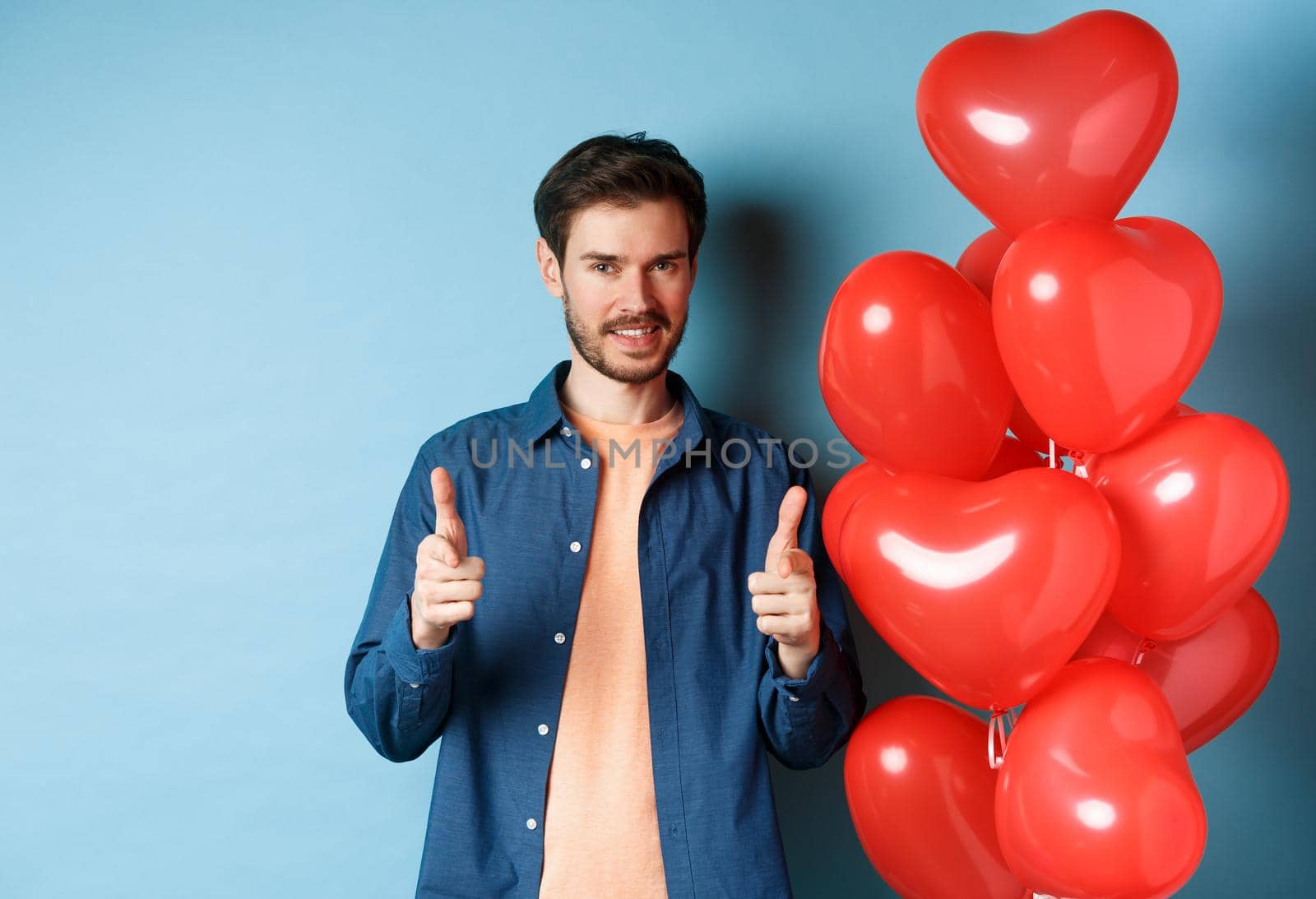 Happy valentines day. Confident man pointing fingers at camera and smiling, standing with balloons on blue background by Benzoix