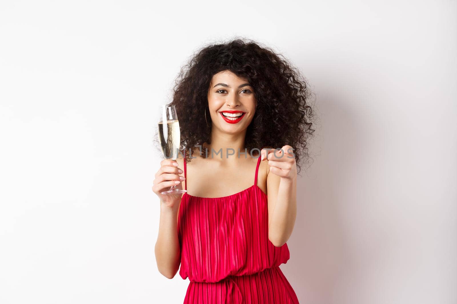 Beautiful curly woman in red dress, partying or having romantic date, holding glass of champagne and pointing at you, inviting person, standing on white background by Benzoix