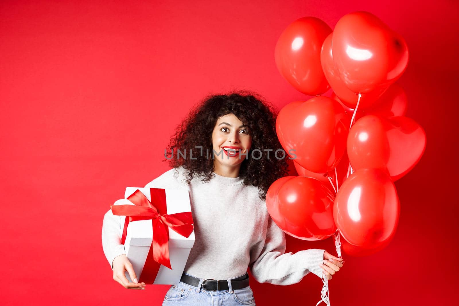 Excited beautiful woman celebrating Valentines day holiday, holding gift box and romantic heart balloons, looking surprised at camera, standing over red background.