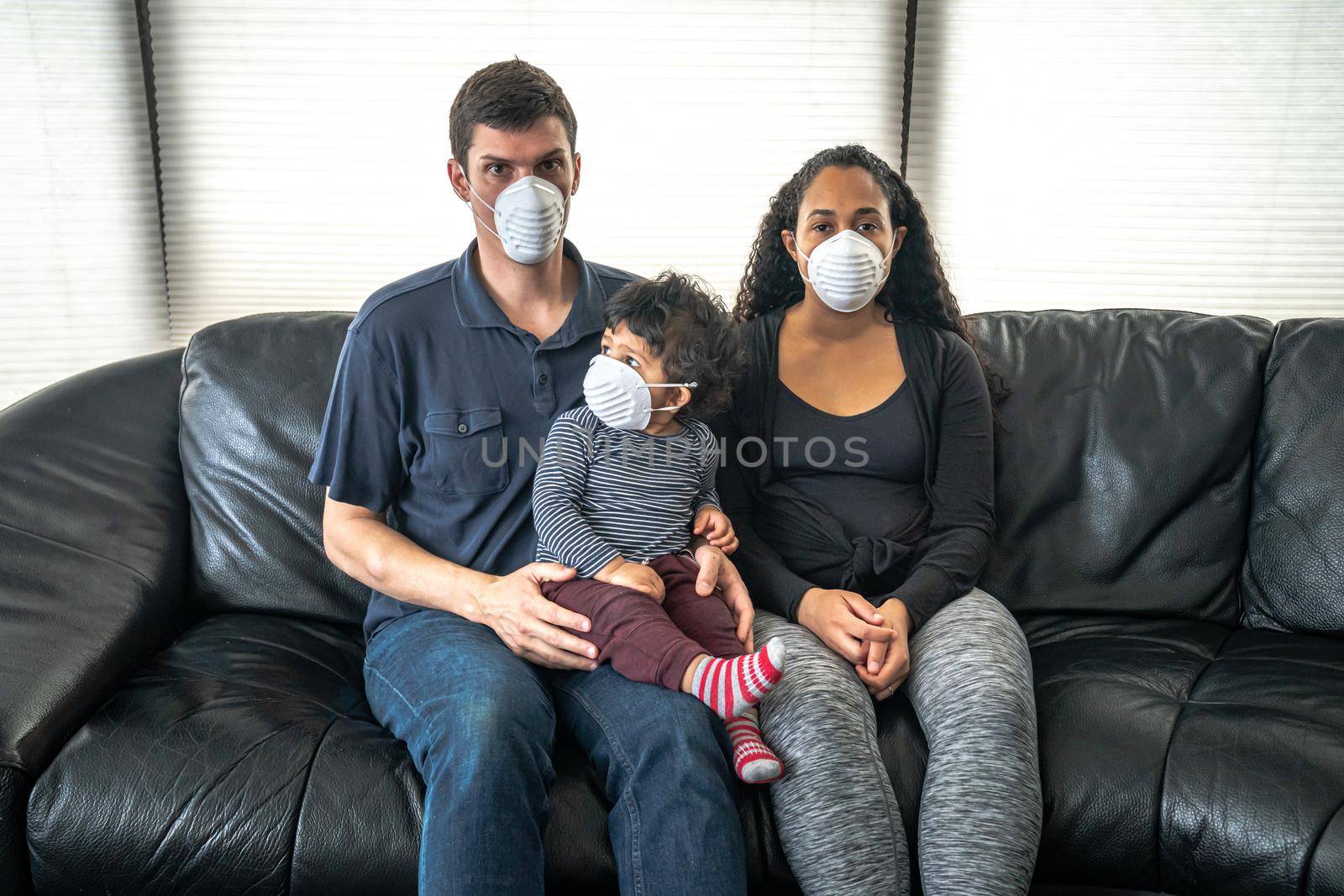 A young mixed family sits on a black leather sofa holding their son and wearing a dust face mask over their faces in hopes of preventing getting sick or ill from caronavirus or COVID-19. by lapse_life