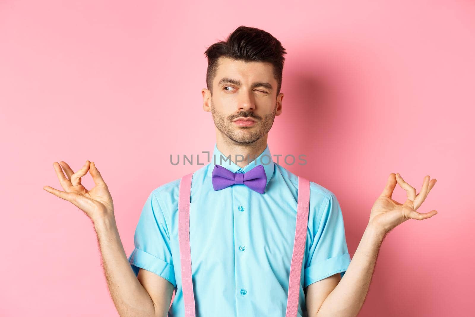 Funny guy with moustache and bow-tie fake meditating, peeking aside while doing yoga asana, standing over pink background.